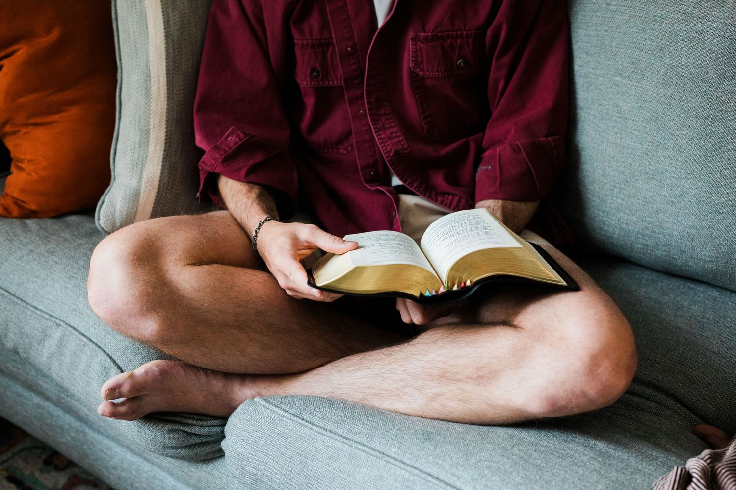 Man sitting cross-legged on a gray sofa reading an open Bible indoors.