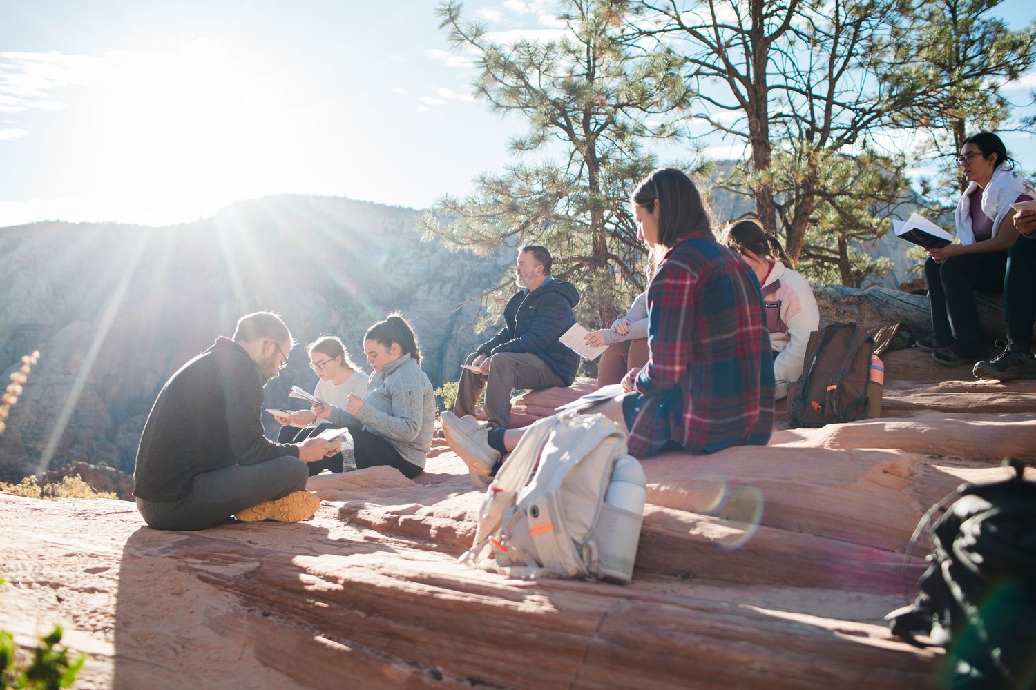 A group of people sitting on red rocks enjoying a sunny afternoon in a mountainous outdoor setting.