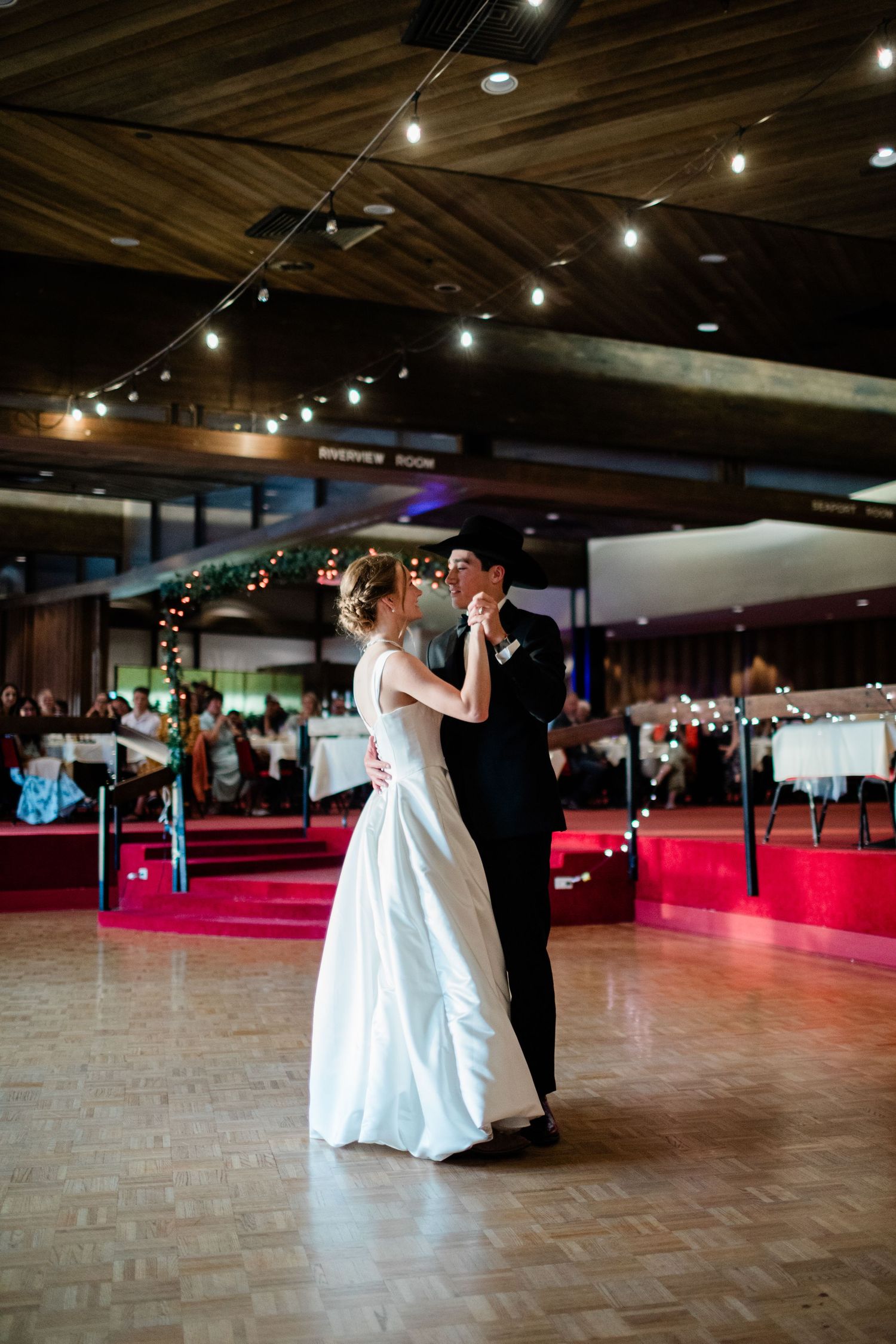 A couple dances together in a spacious ballroom with string lights overhead and a red-lit dance floor.