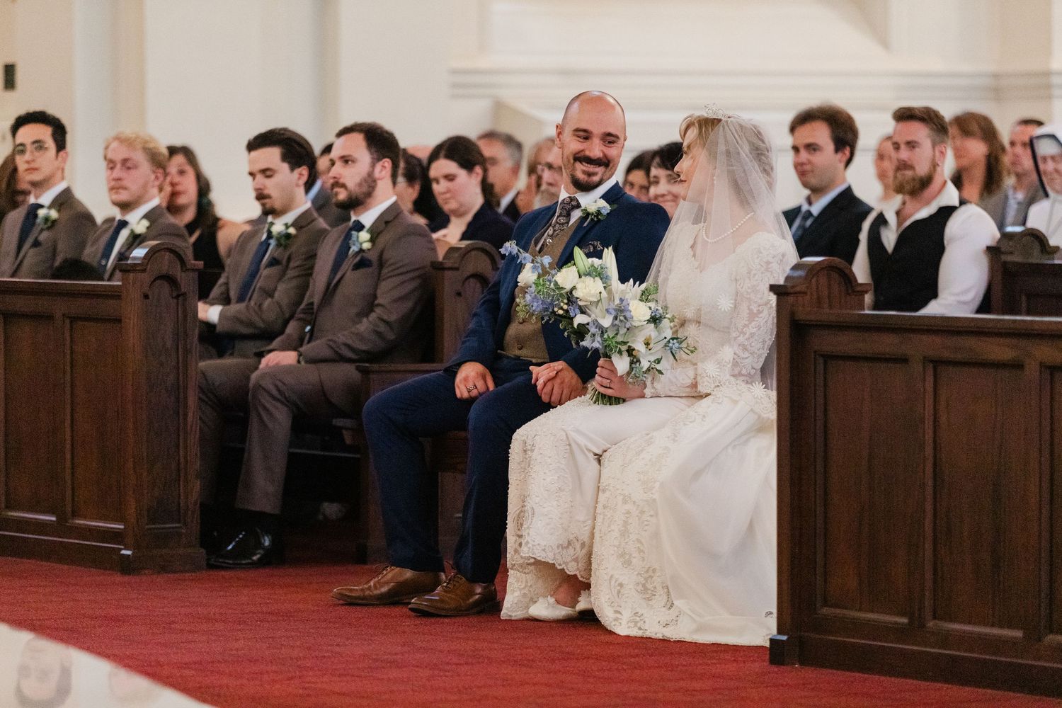 Bride and groom sit together in a church pew during their wedding ceremony surrounded by guests.