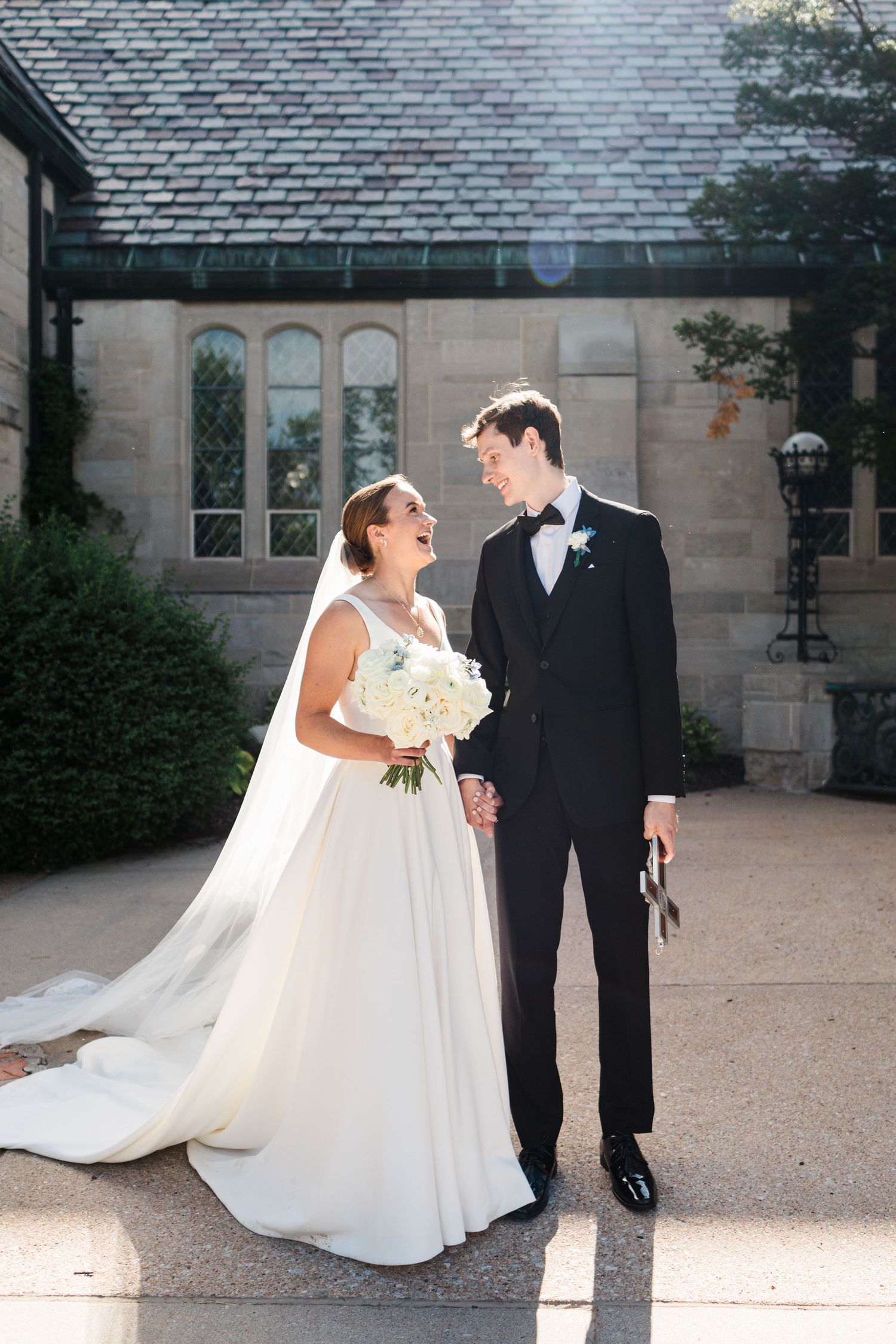Bride and groom laughing together outside a stone chapel, bride holding white bouquet in elegant ball gown and veil.