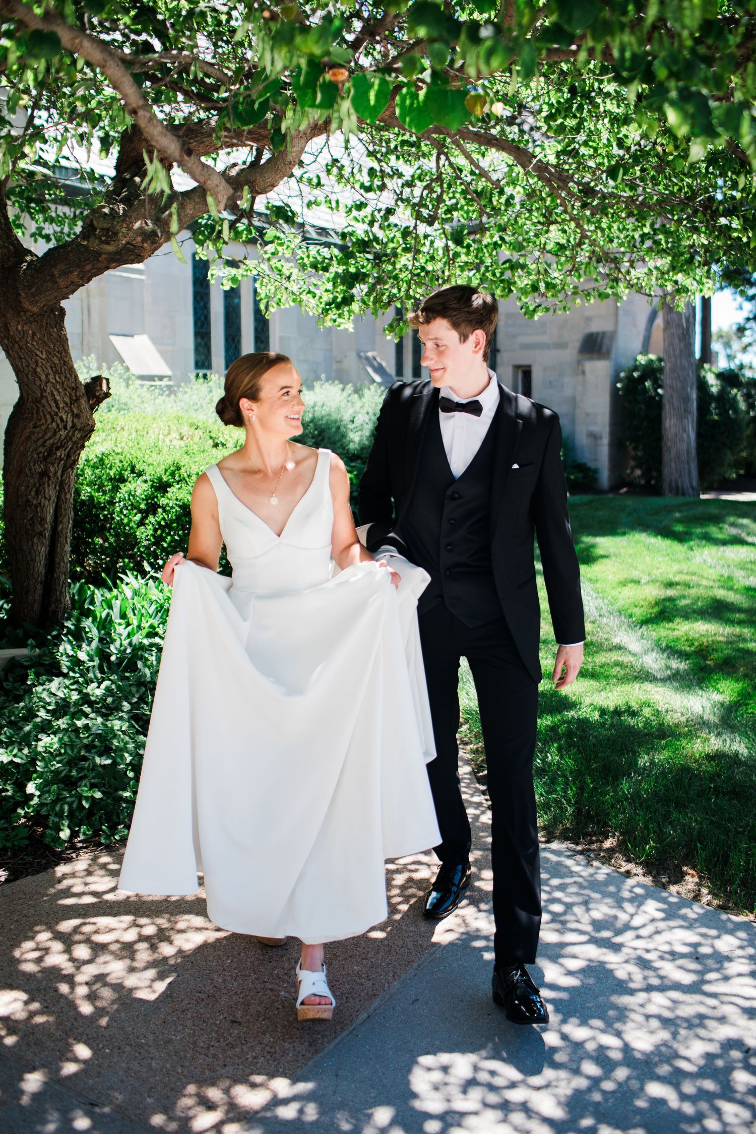 Bride in simple white gown and groom in black tuxedo walk together under lush green tree outdoors.