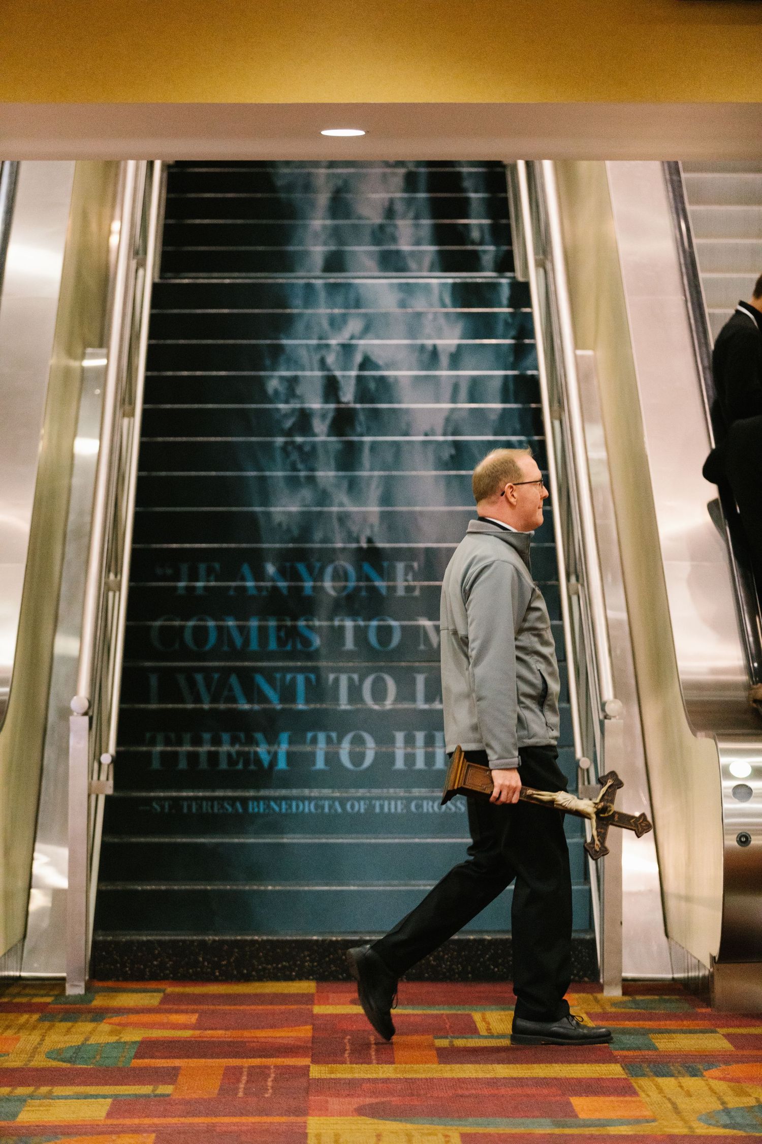 People ascending illuminated escalator in a convention center with colorful patterned carpet.