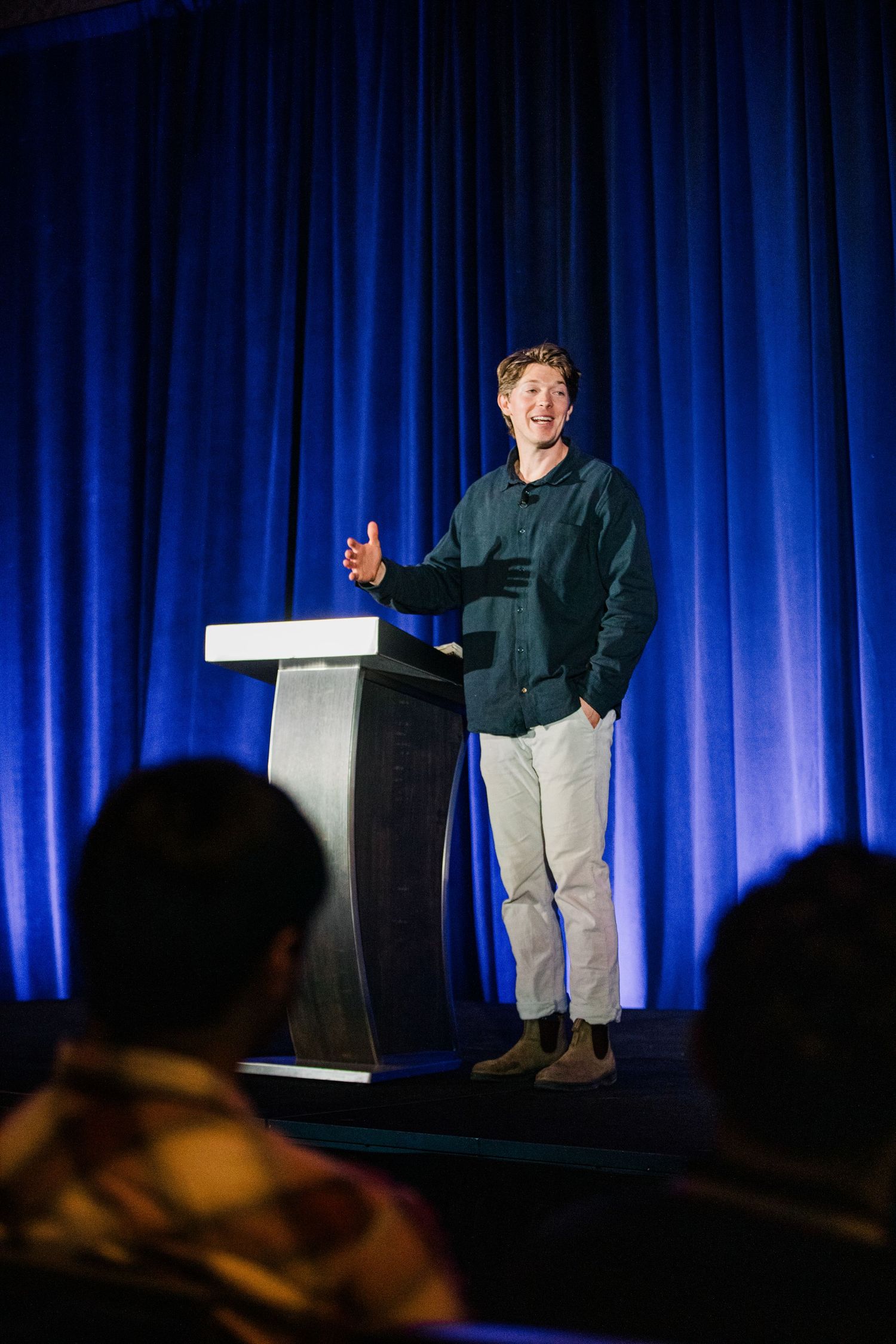A person stands on a stage with blue curtains and dramatic lighting during a presentation.