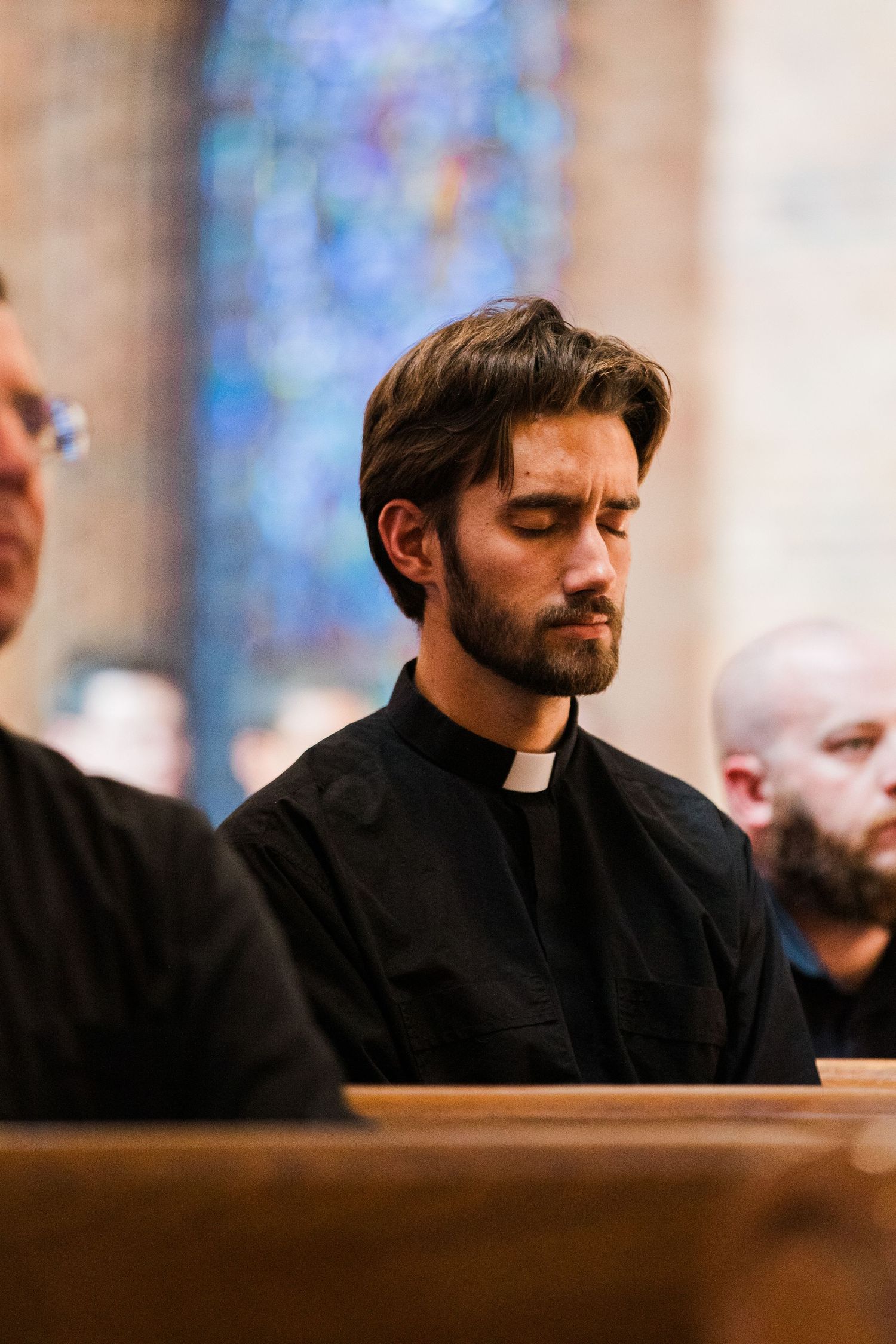 Young priest in black clerical collar praying with eyes closed in church pew with stained glass background.