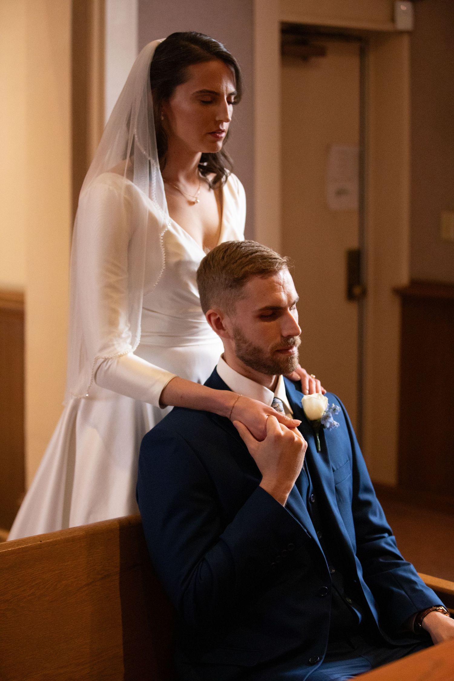 Bride in white gown and veil holds groom's hand as he sits in church pew wearing navy suit with boutonniere.