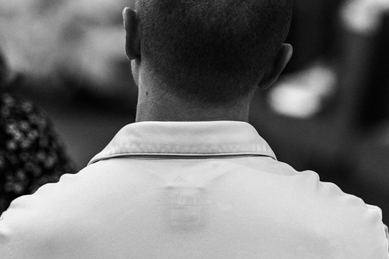 Black and white photo of a man seen from behind, showing his shaved head and white collared shirt.