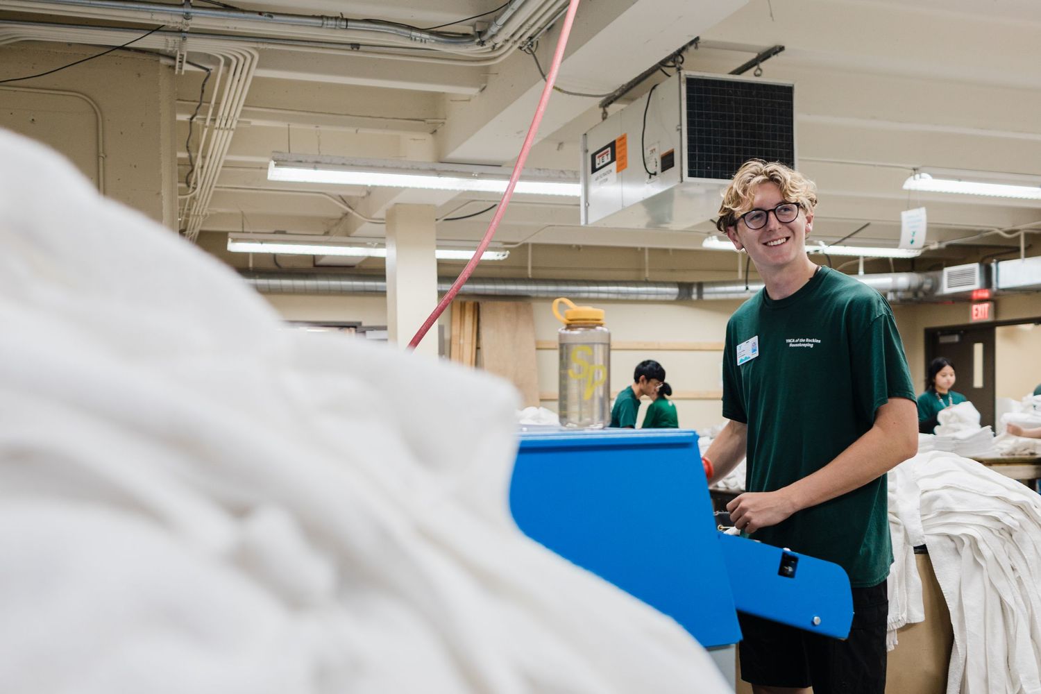 Young man in green shirt smiling while working in a commercial laundry facility surrounded by white linens.