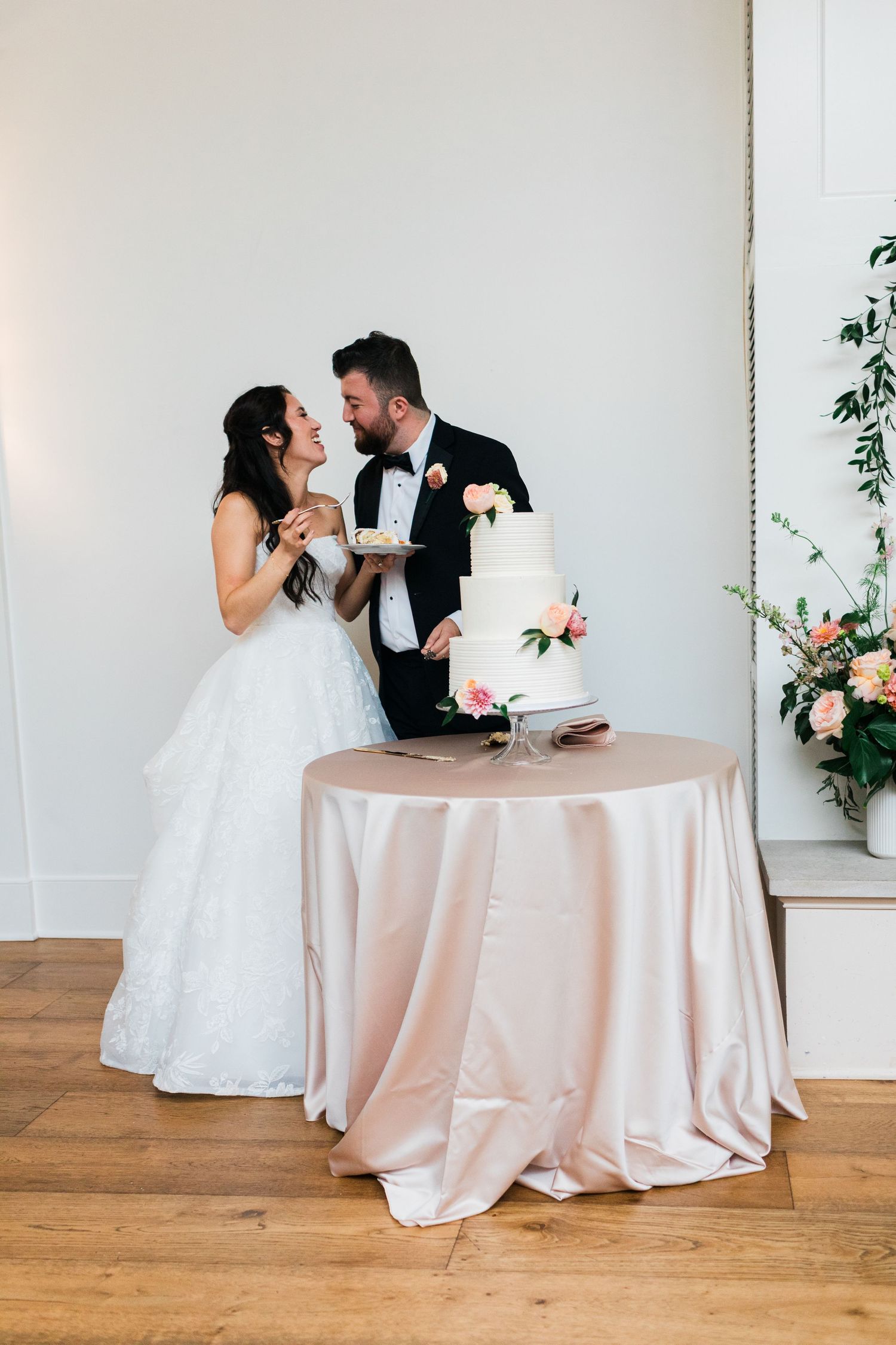 Bride and groom cutting a white tiered wedding cake on a blush pink tablecloth in an elegant venue.