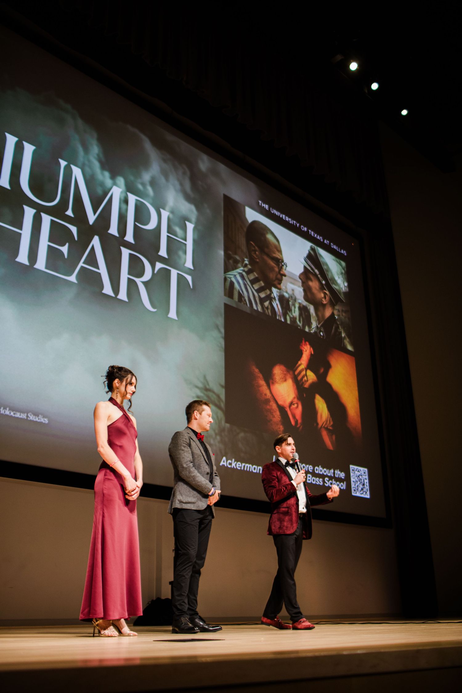 Three performers stand on stage at a film screening with 'Triumph Heart' displayed on the projection screen behind them.