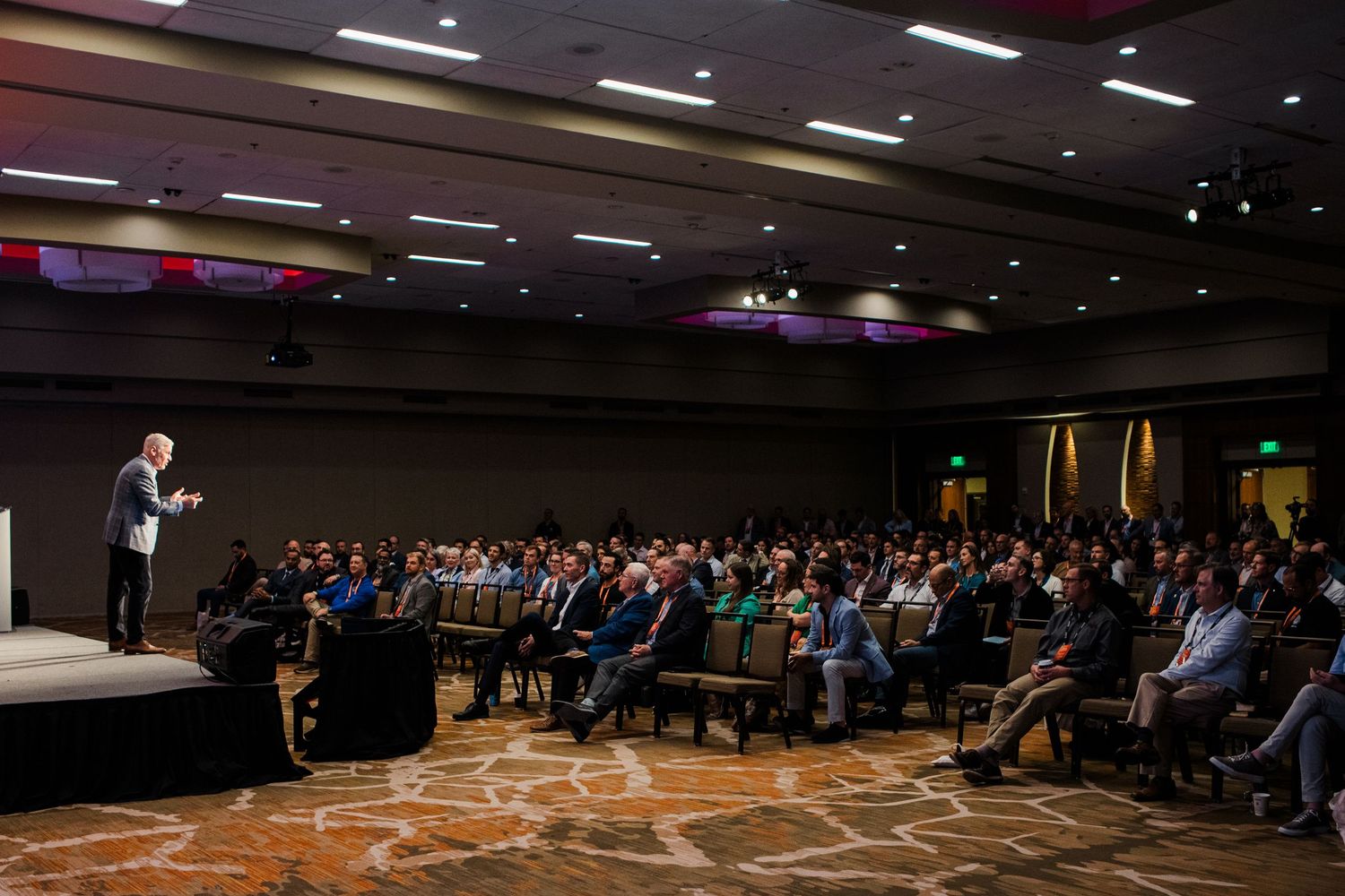 A keynote speaker addresses a large audience in a packed conference ballroom during a professional event.