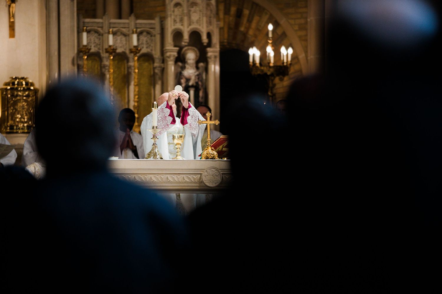 Priest performing Catholic Mass at ornate altar with candles, congregation silhouetted in foreground of grand church.