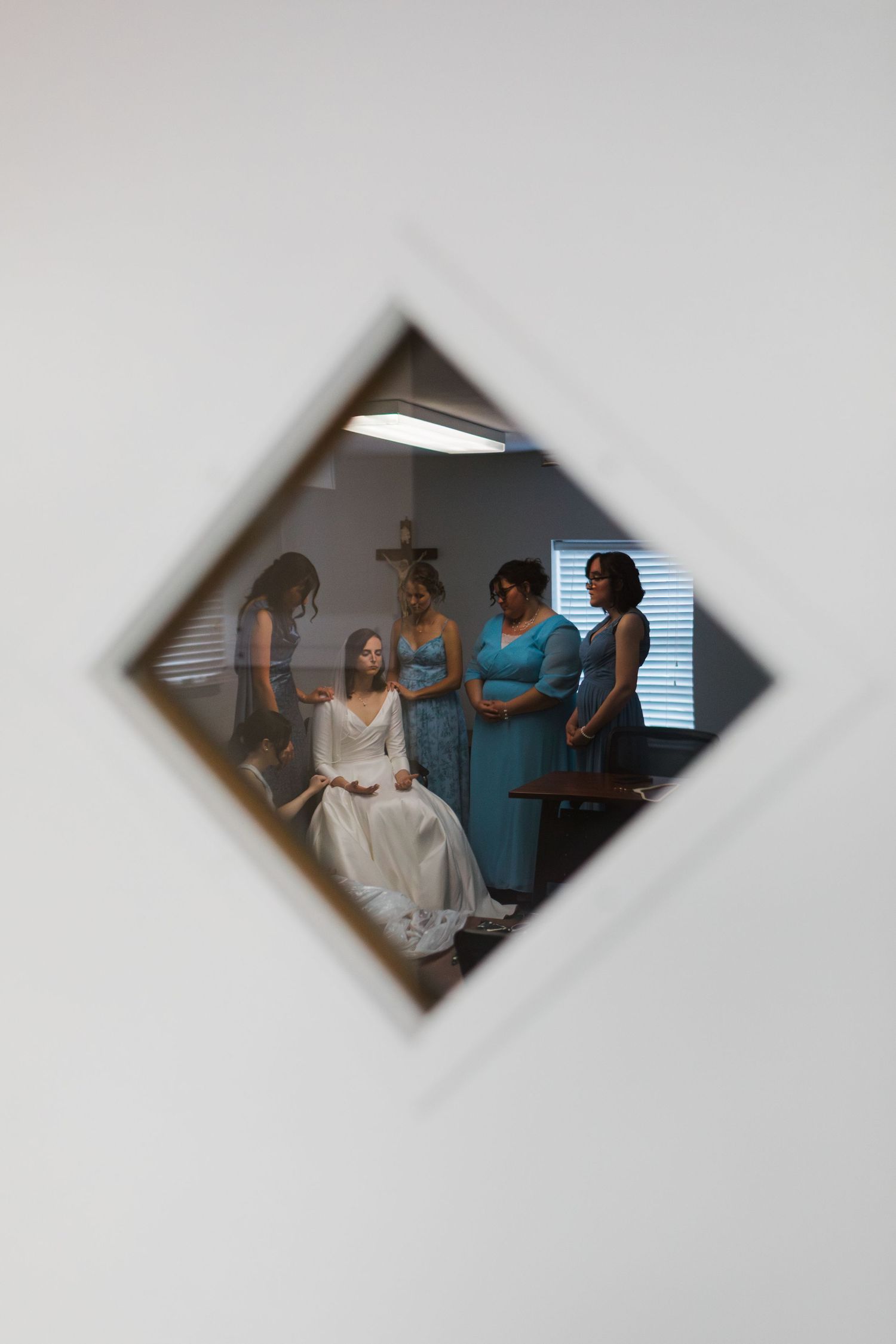 Bride in white gown seated as bridesmaids in teal dresses assist her, reflected in a diamond-shaped mirror.