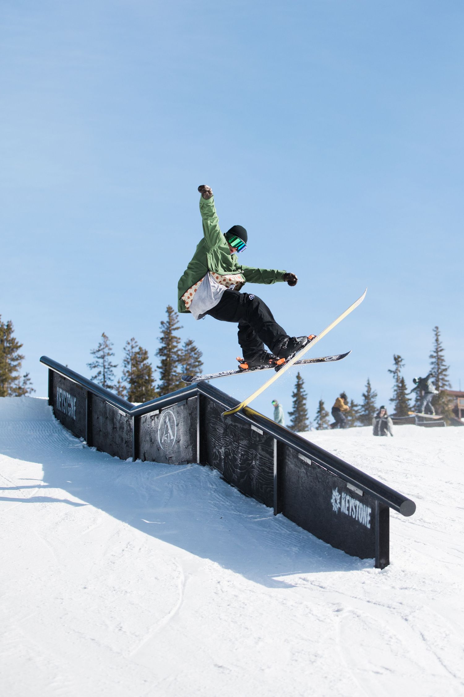 A snowboarder executes an aerial trick over a black rail feature in a terrain park with evergreen trees.