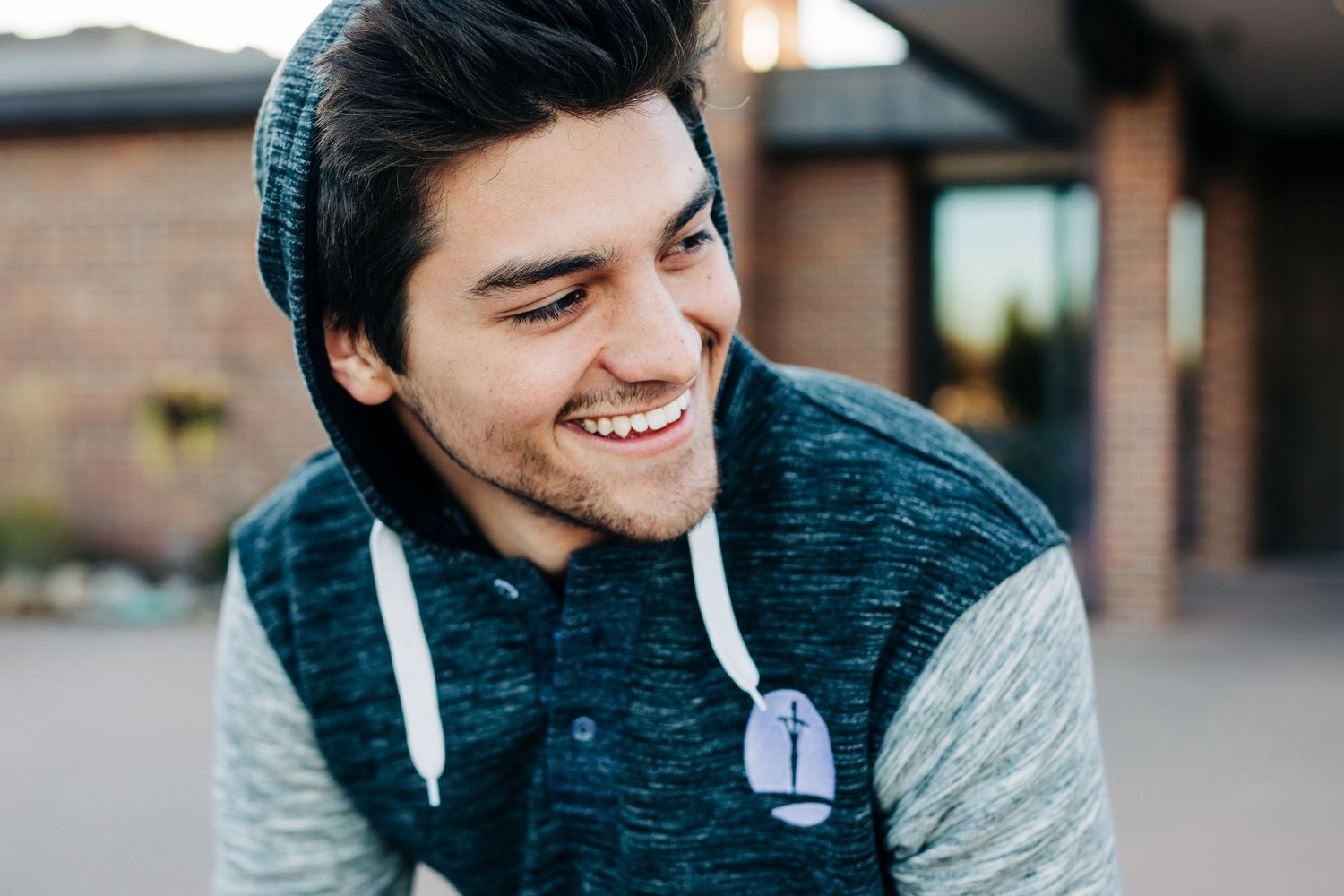 A young man in a two-toned hoodie smiles warmly while looking down during a casual outdoor portrait session.