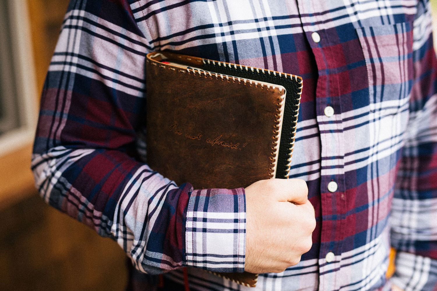 Person wearing plaid flannel shirt holds brown leather wallet close to their chest.