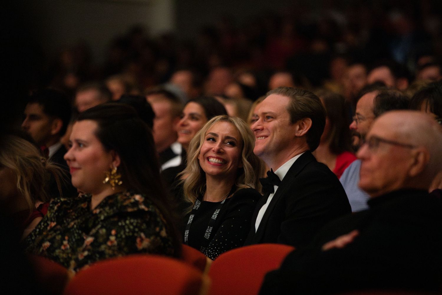Audience members enjoy a performance in a dark theater with red seats.