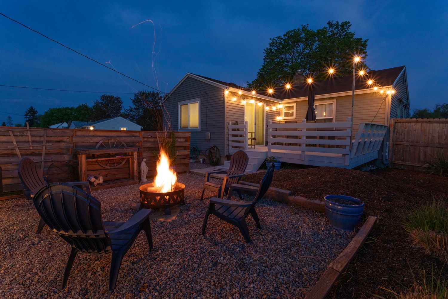 A cozy outdoor seating area with a fire pit, surrounded by wooden fencing and illuminated by string lights at dusk.
