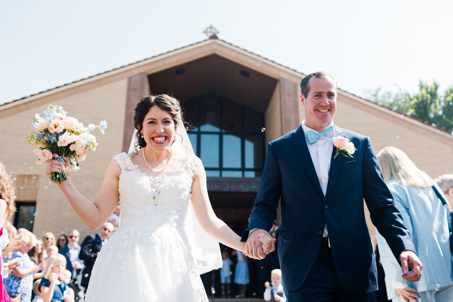 Happy newlyweds exit church holding hands, bride in lace gown with bouquet, groom in navy suit with bow tie.