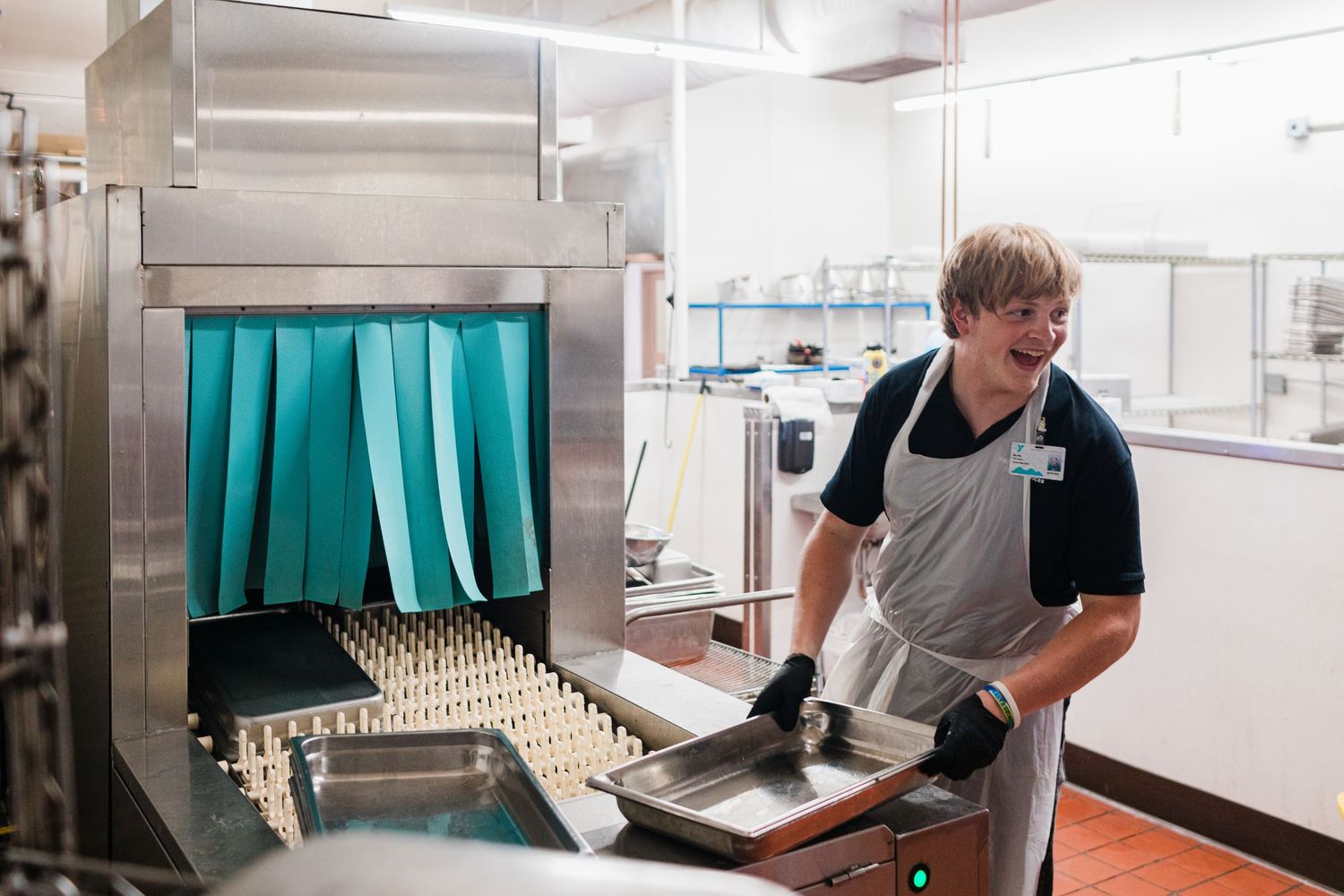 A smiling kitchen worker in an apron loads dishes into a commercial stainless steel dishwasher machine.