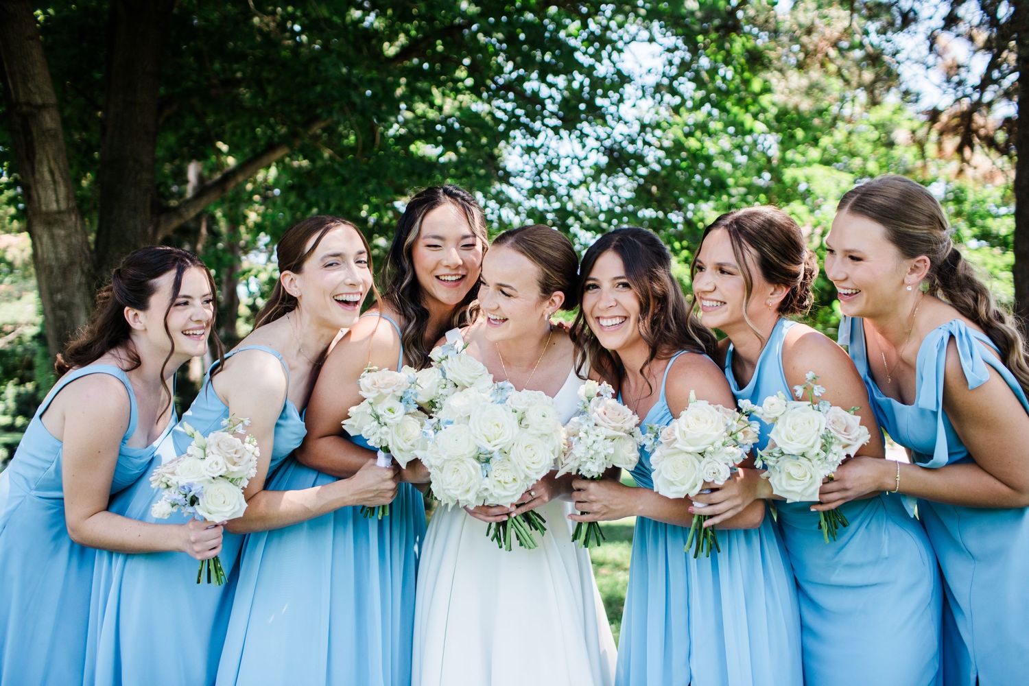 Bride and bridesmaids in light blue dresses laughing together outdoors holding white bouquets.
