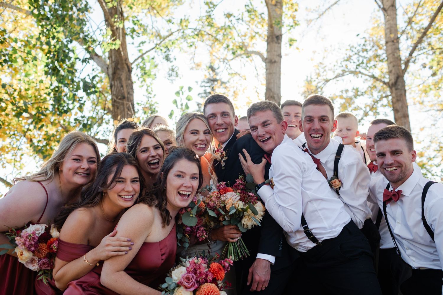 Wedding party group photo with bridesmaids in burgundy dresses and groomsmen in white shirts, smiling outdoors among autumn trees.
