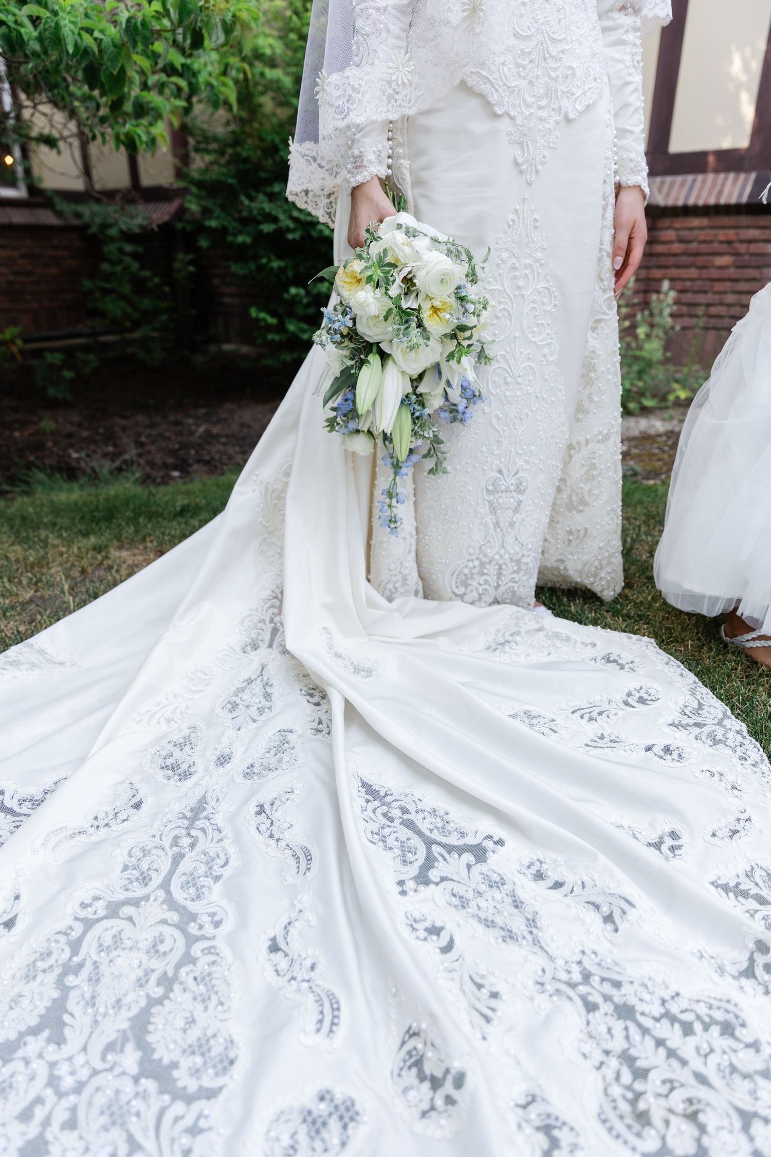 Close-up of a brides intricate lace wedding gown train spread on green grass with floral bouquet.