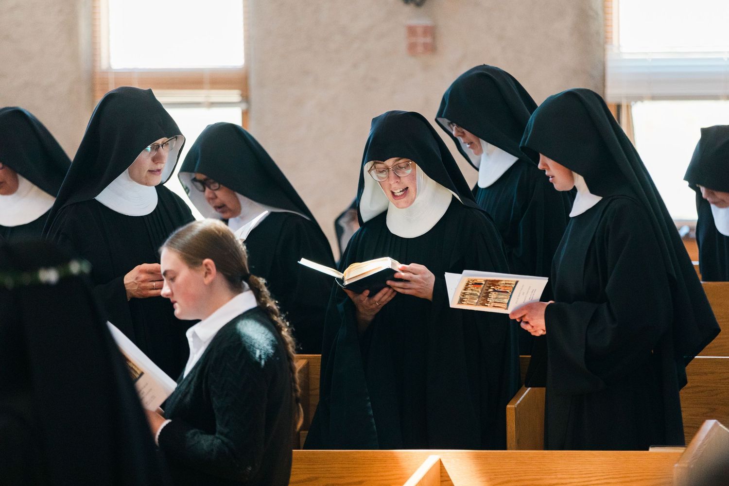 A group of nuns in black habits read books and converse together in church pews.
