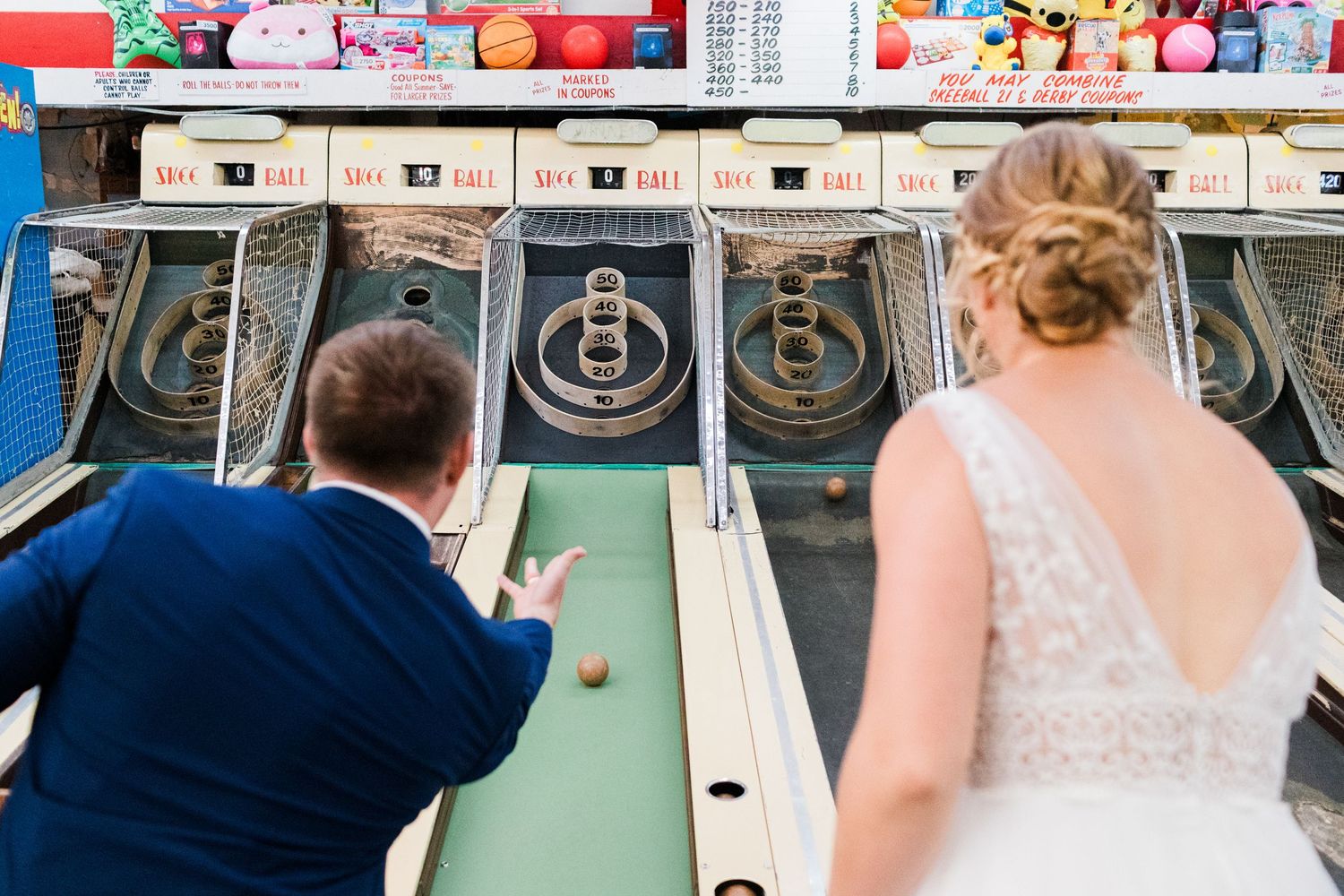 Bride and groom play skee-ball together at a fun arcade during their unique wedding reception.