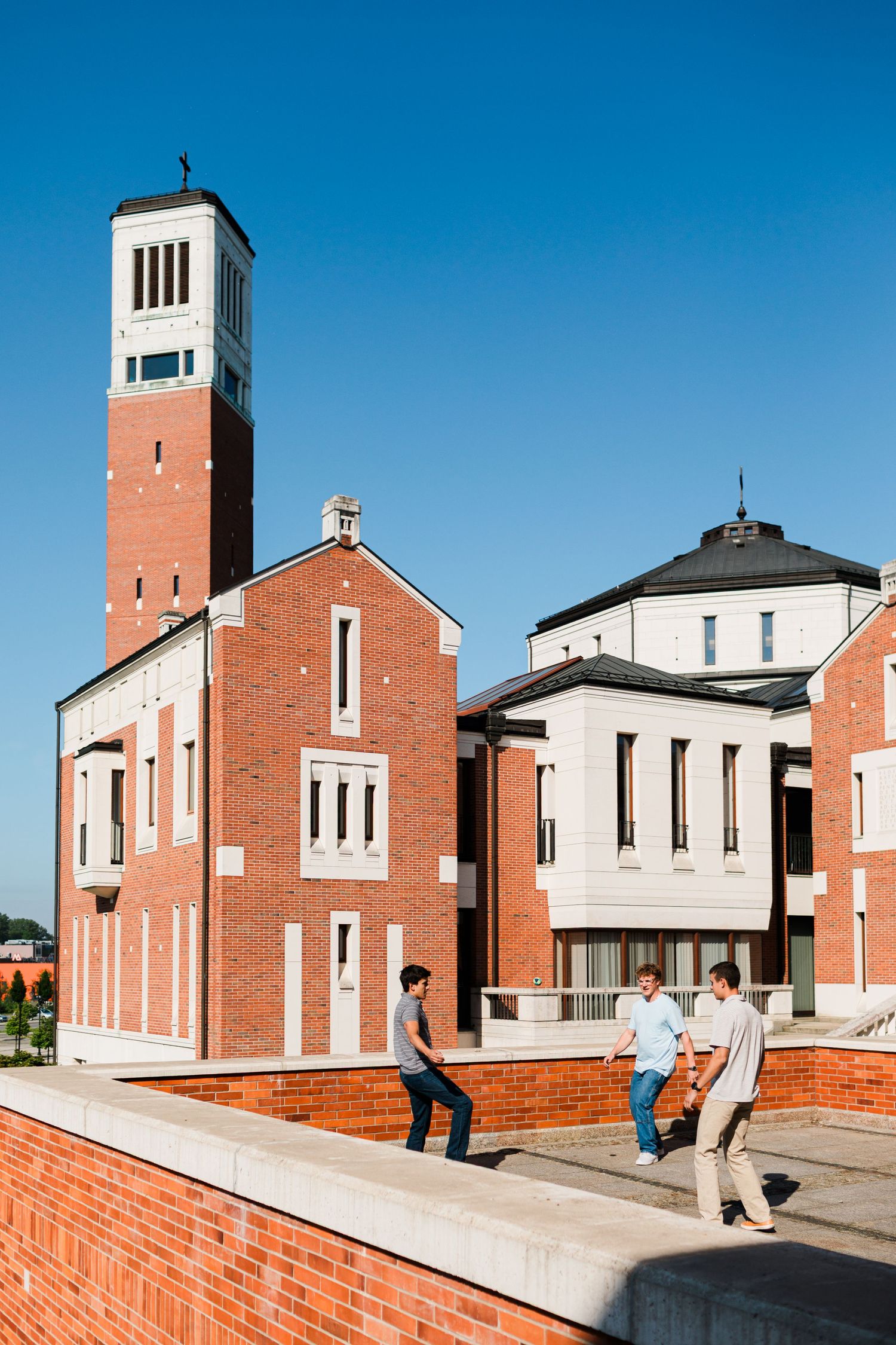 Red brick church with tall bell tower and modern dome building under clear blue sky with three people walking nearby.