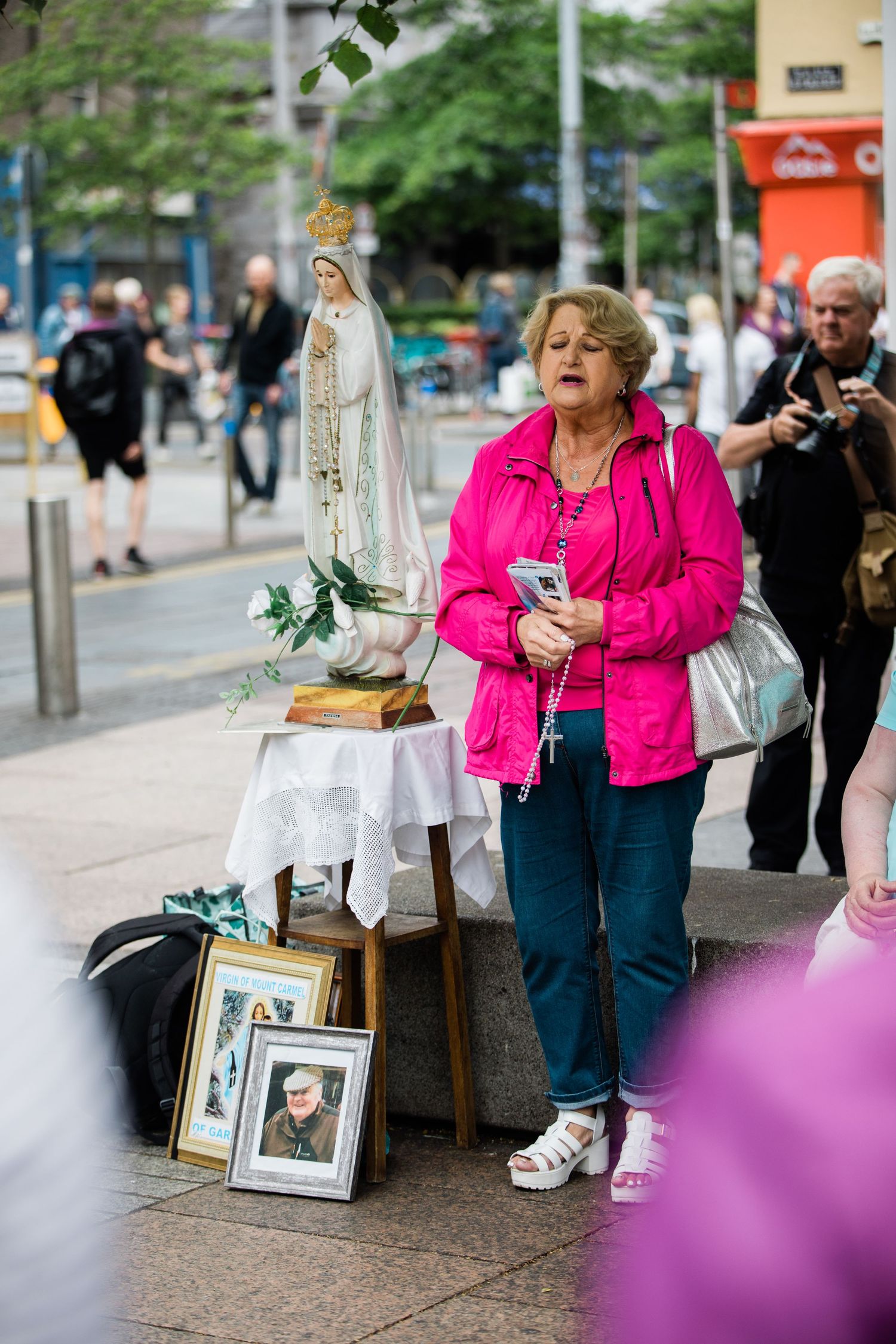 Woman in pink jacket praying with rosary beside a Virgin Mary statue on a city sidewalk.
