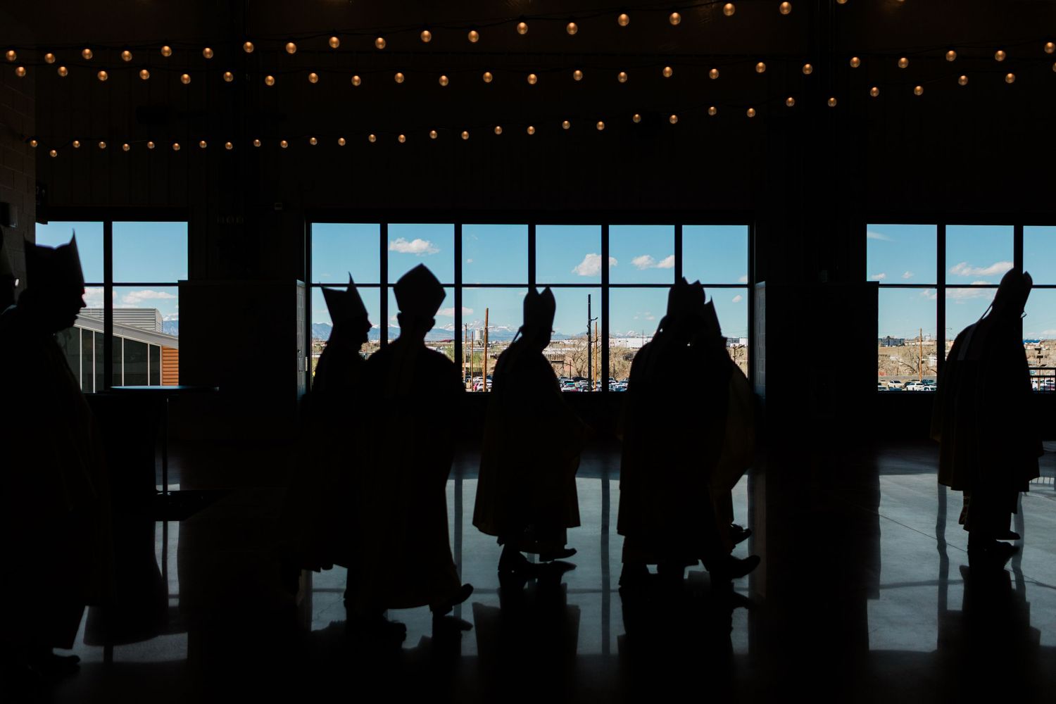 Silhouettes of Catholic bishops in miters processing through a grand hall with large windows.