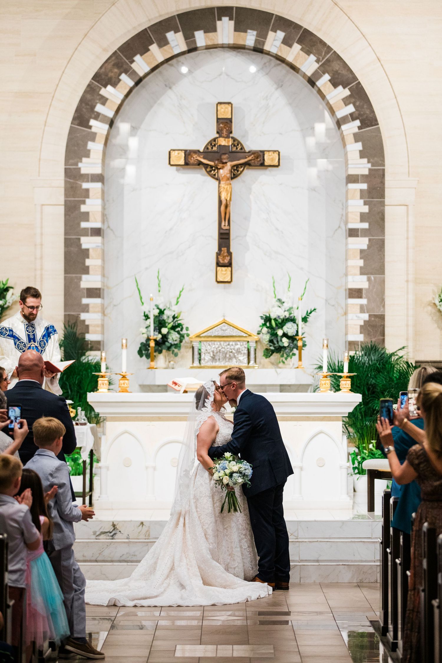 Bride and groom share first kiss at Catholic church altar with crucifix backdrop during wedding ceremony.