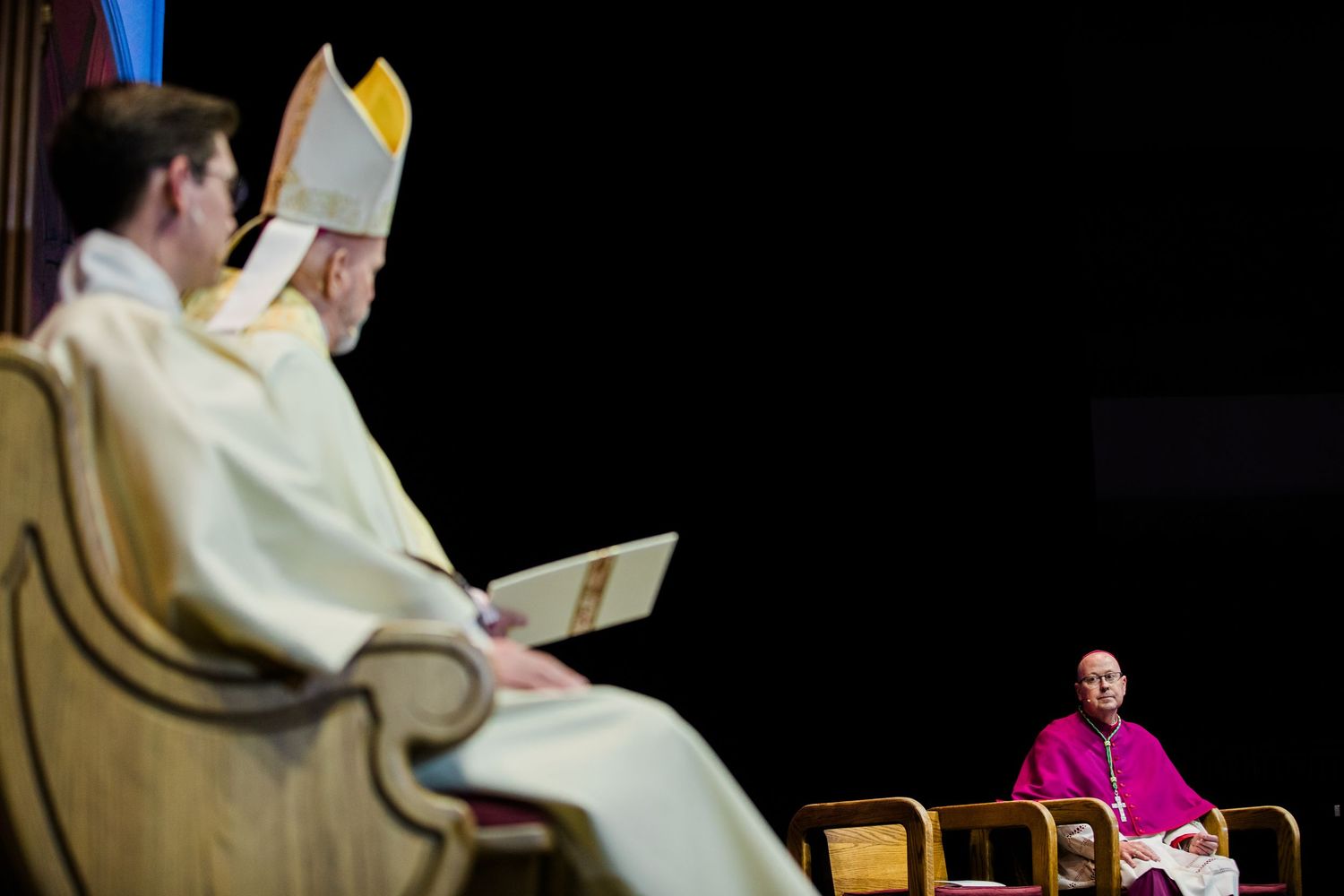 Catholic clergy members seated in ornate church pews during a religious ceremony with colorful lighting.