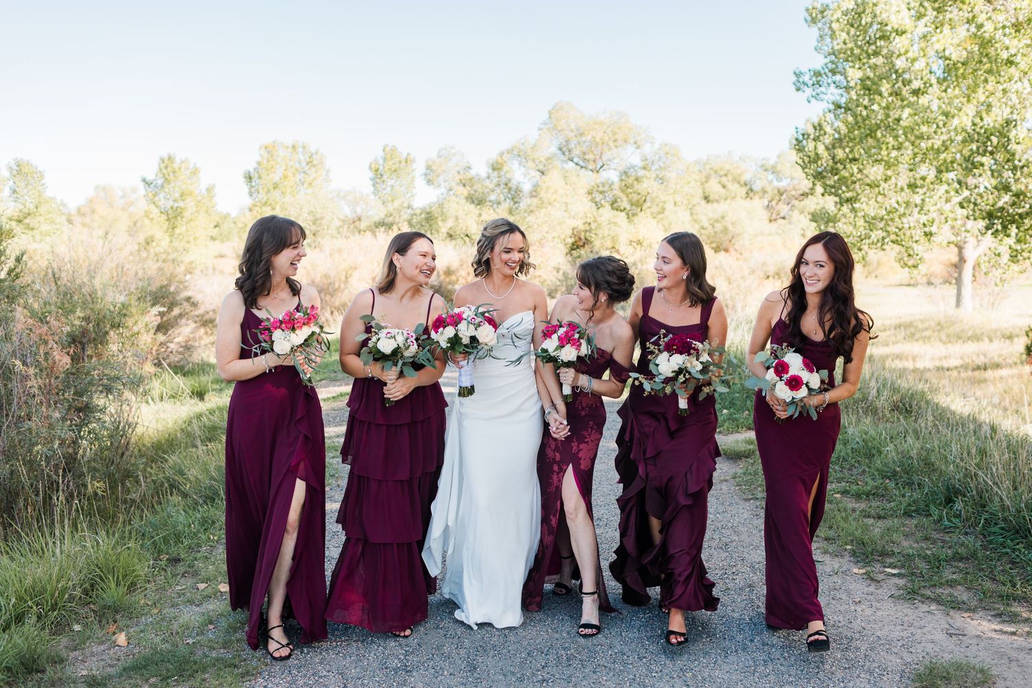 Bride in white gown walks with five bridesmaids in burgundy dresses, holding bouquets on a sunny outdoor path.