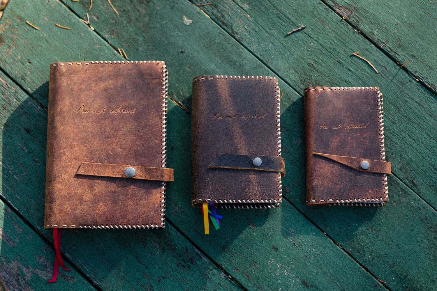 Three handmade leather notebooks with button closures arranged in descending sizes on a weathered green wooden surface.