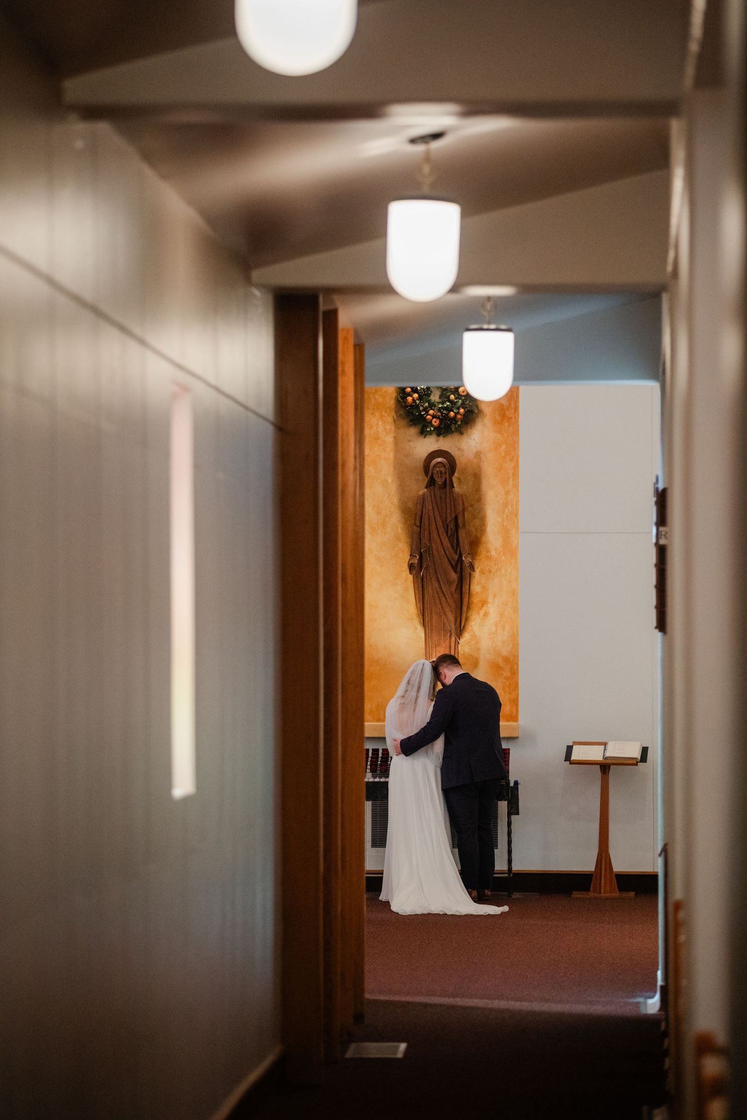 A hallway with white walls and wooden doors leads to a distant figure in white and black formal attire.