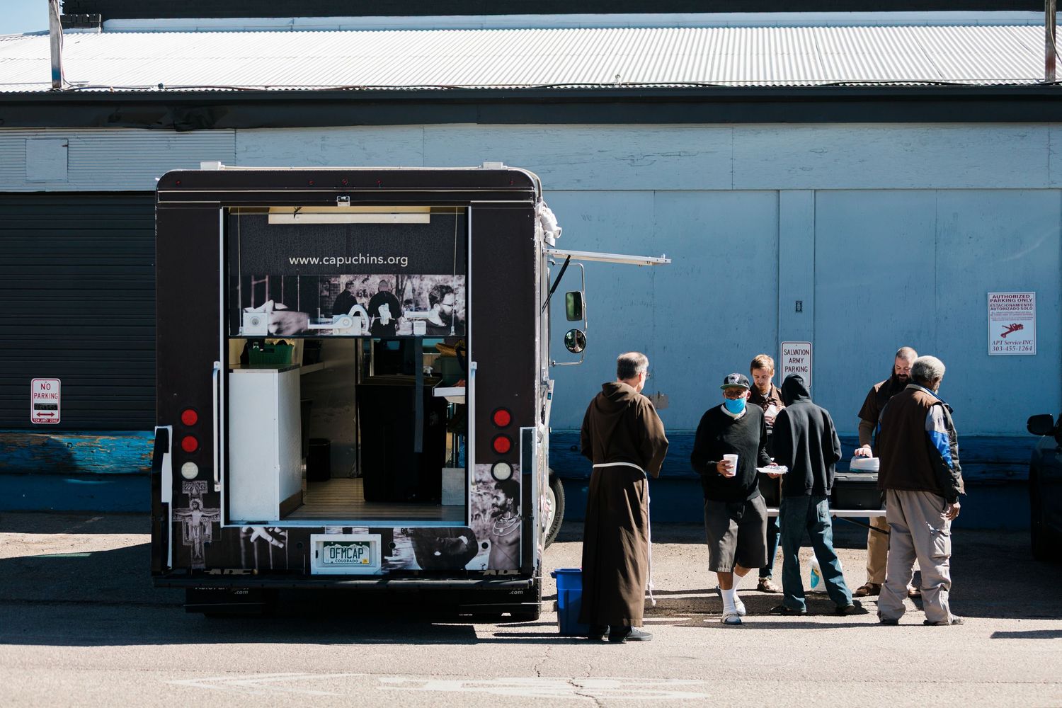 People gather outside a black food truck parked against a blue warehouse wall on a sunny day.