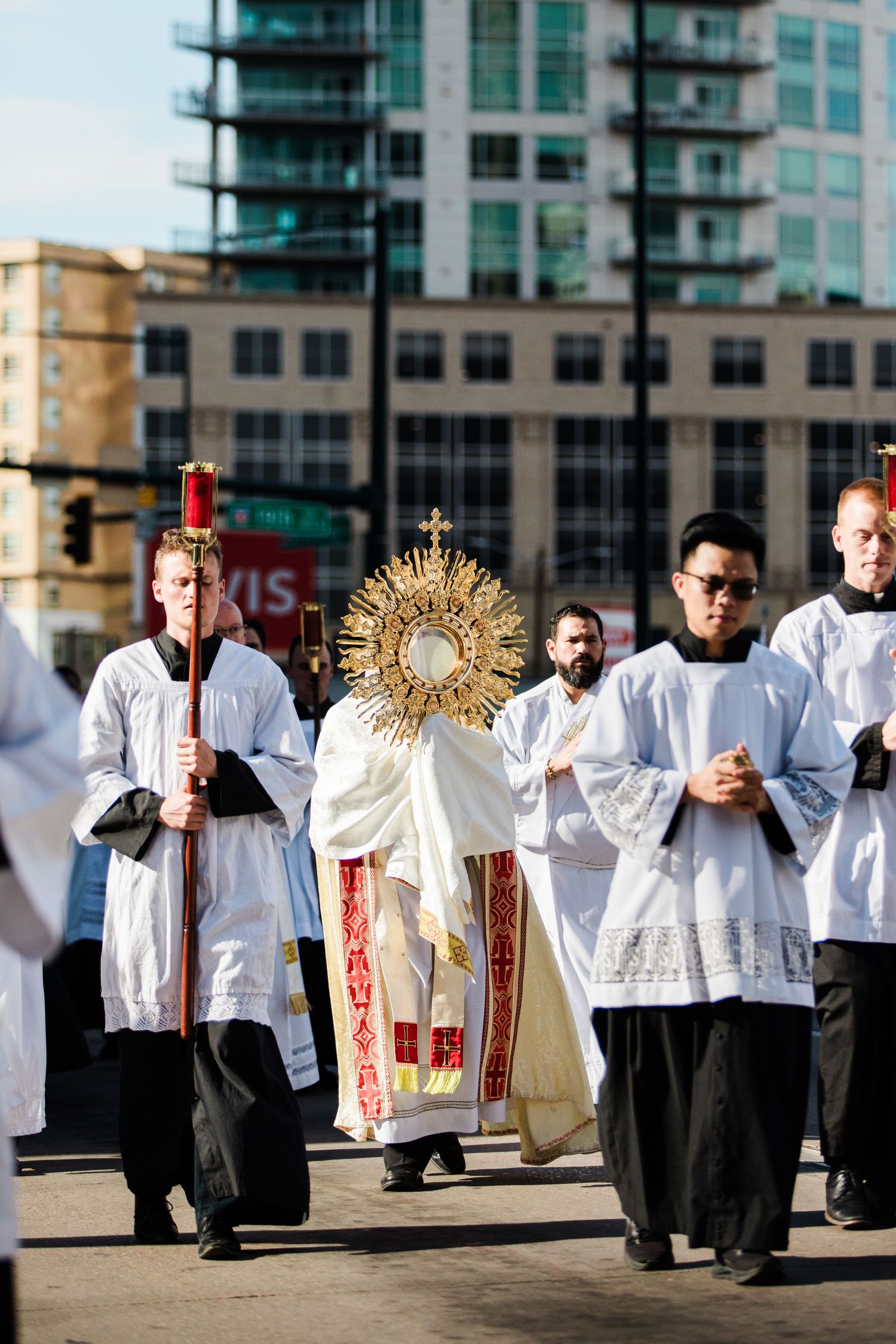 Catholic clergy carry a golden monstrance in a Corpus Christi procession through an urban street.
