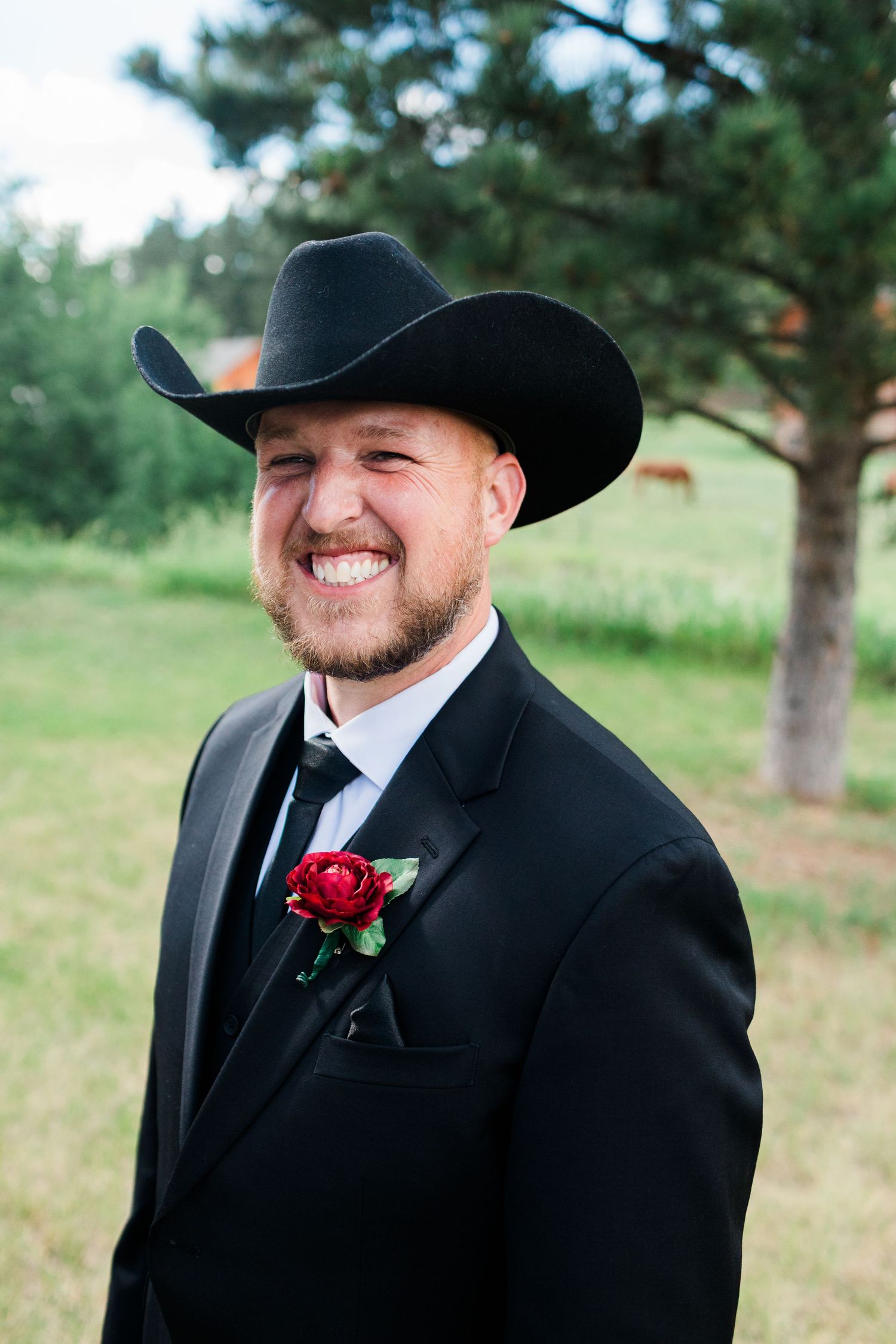 Smiling groom in black tuxedo and cowboy hat with red rose boutonniere at outdoor country wedding.