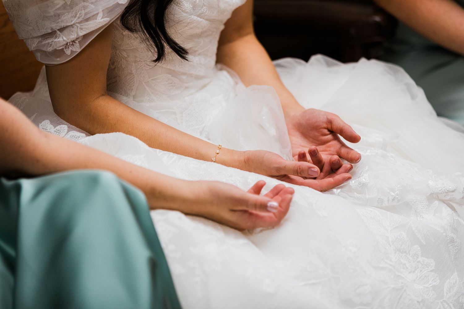 Bridesmaid in sage green dress holds bride's hands over white lace wedding gown.