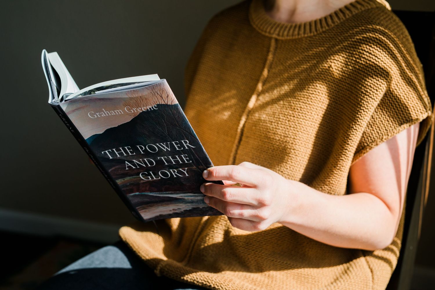 Woman in mustard knit vest reading The Power and the Glory book in warm sunlight.
