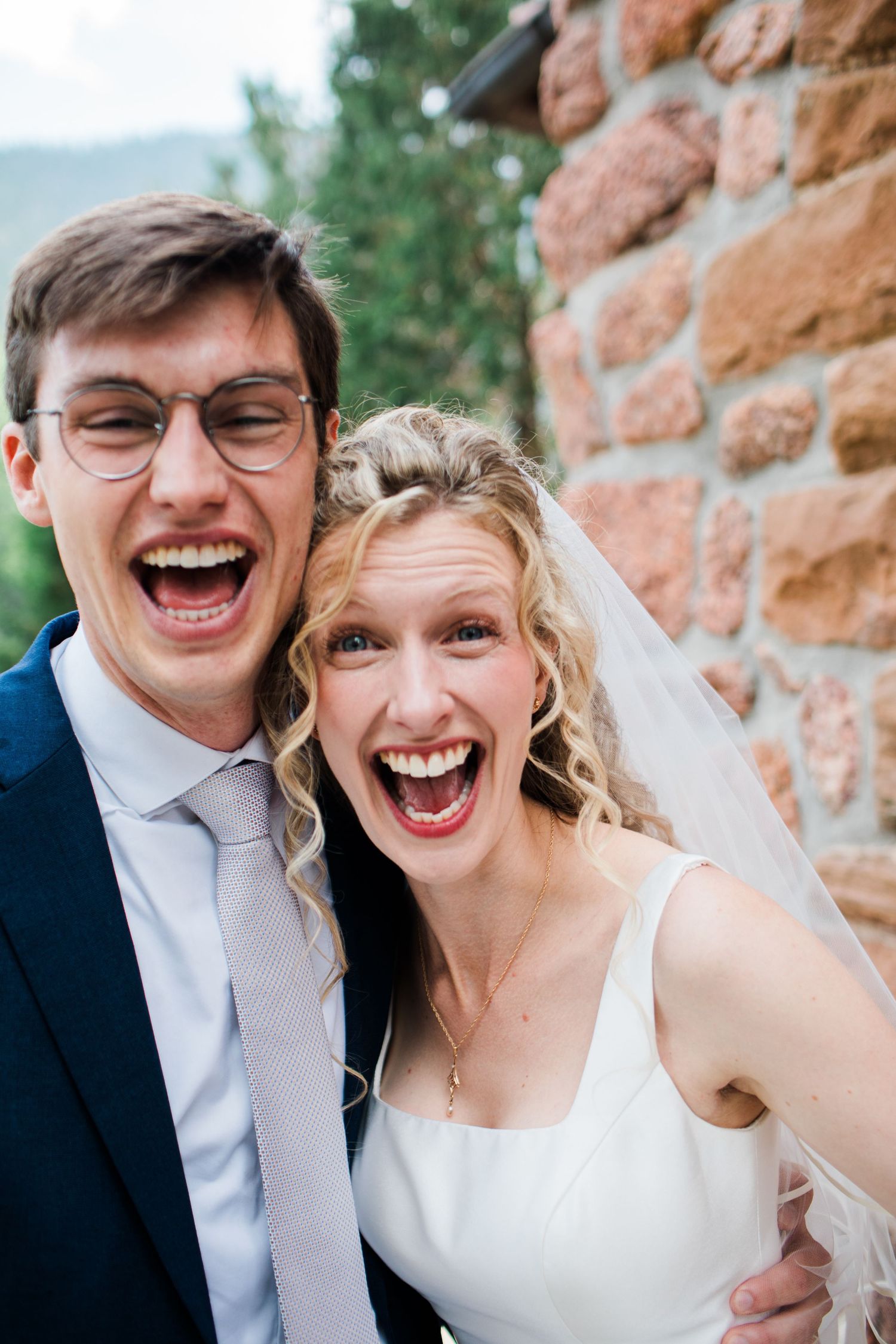 Joyful bride and groom laughing together outdoors near a brick wall, bride wearing a white dress and veil.