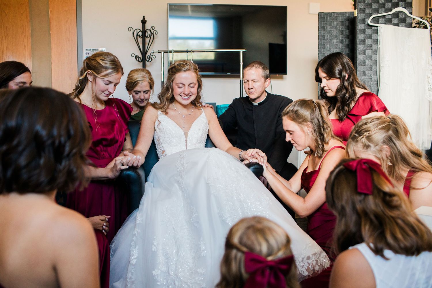 Bride in white gown surrounded by bridesmaids in red dresses helping her prepare on her wedding day.