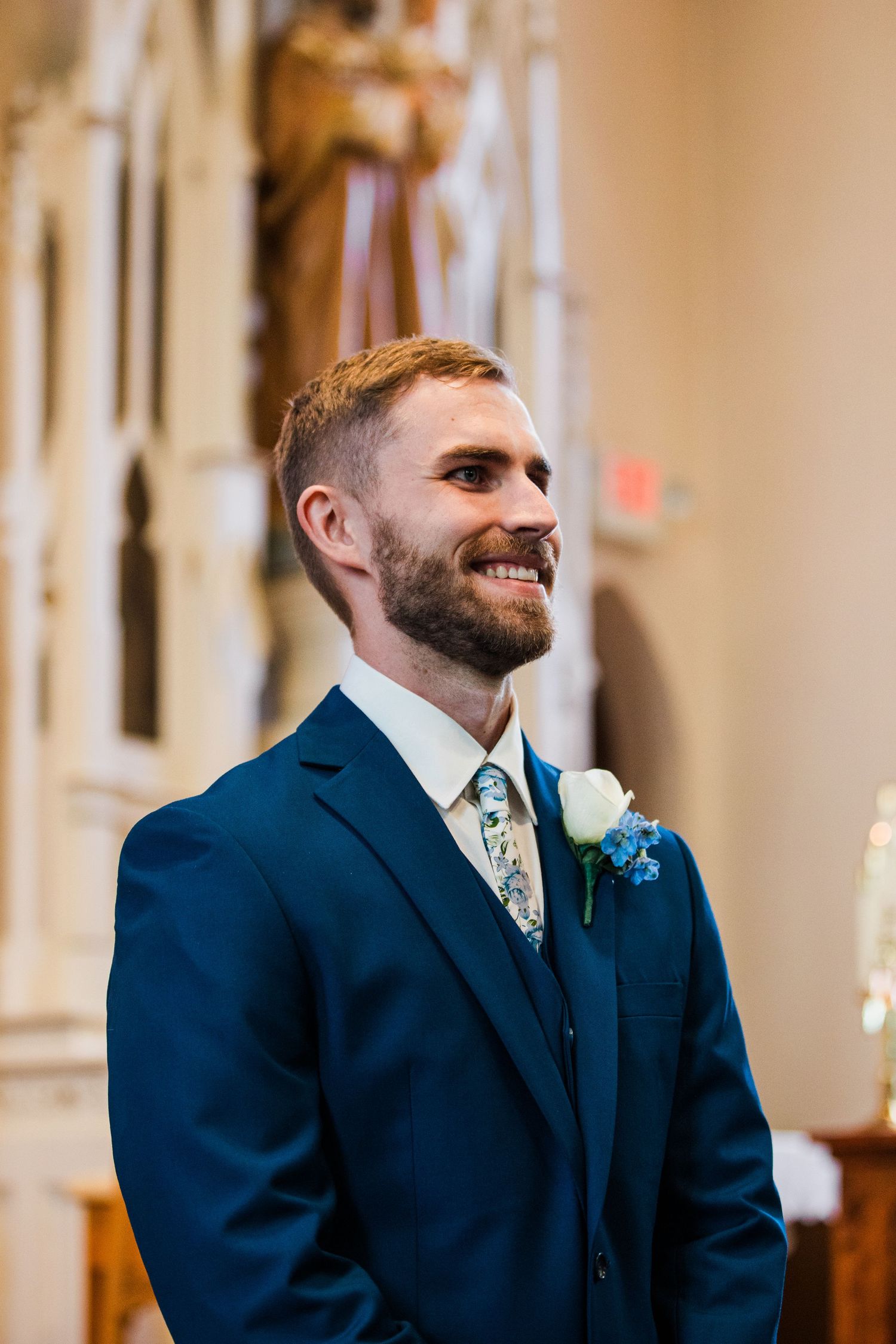 Smiling groom in navy blue suit with floral tie and white boutonniere standing at church altar on wedding day.