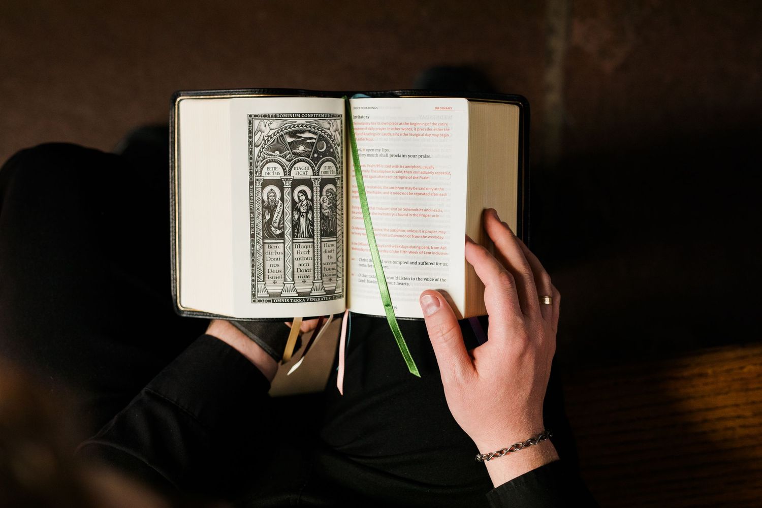 Person reading an open book with religious illustration and green ribbon bookmark, hands visible with bracelet.