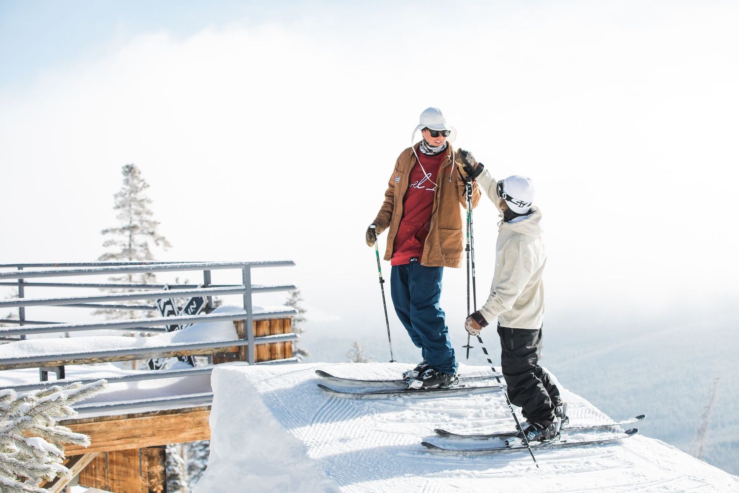 Two skiers enjoy a snowy day at a mountain resort deck with benches and scenic winter views.