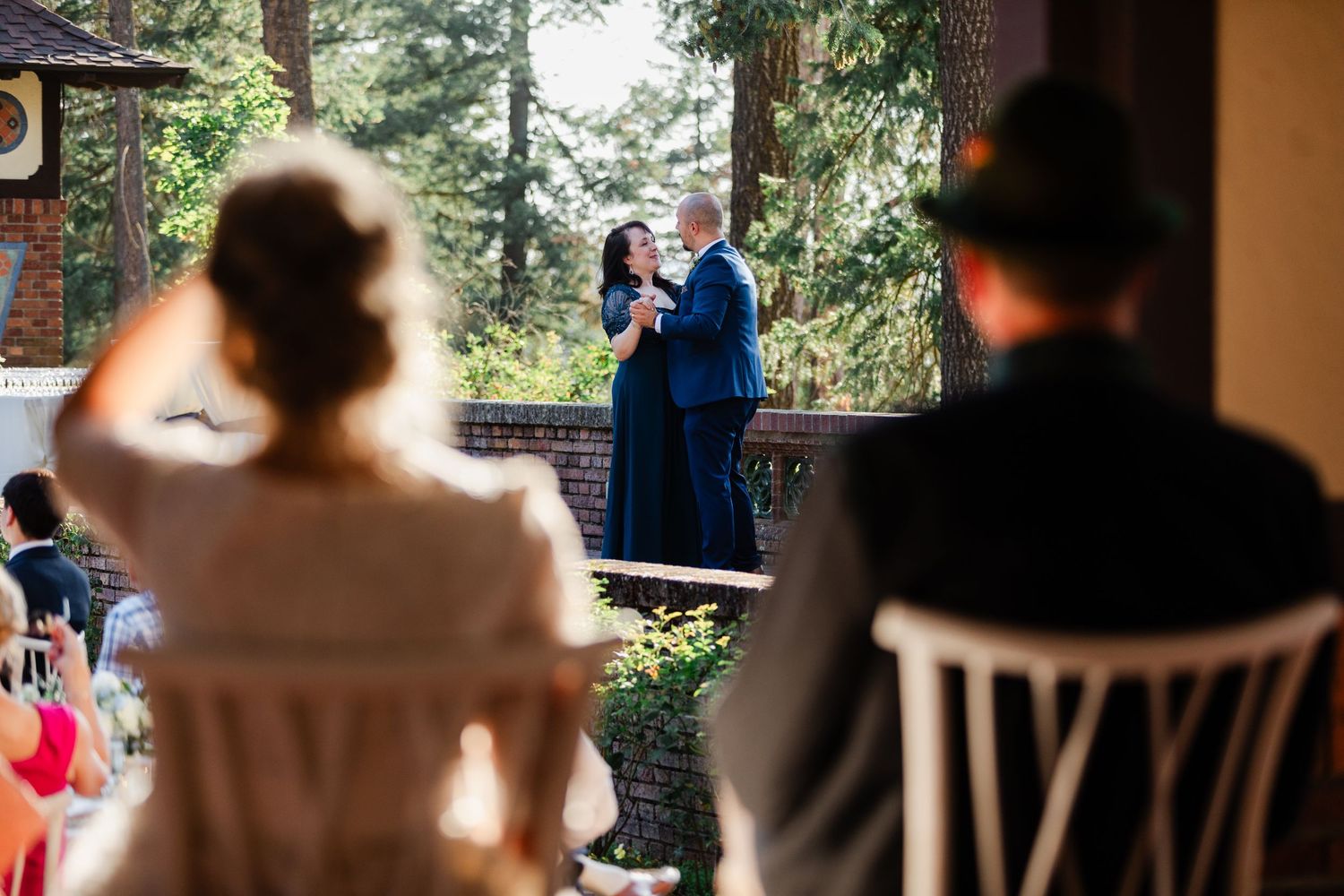 Couple shares first dance on outdoor terrace while wedding guests watch from white chairs at reception tables.