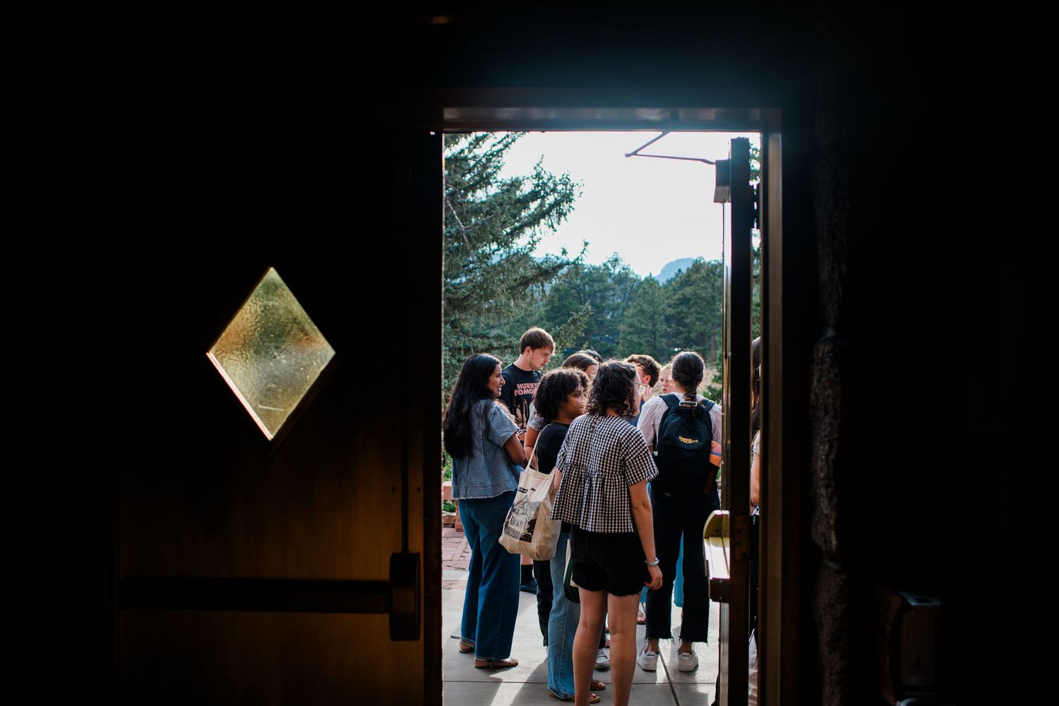 Group of people gathered outside a rustic barn doorway surrounded by trees and mountains in natural light.