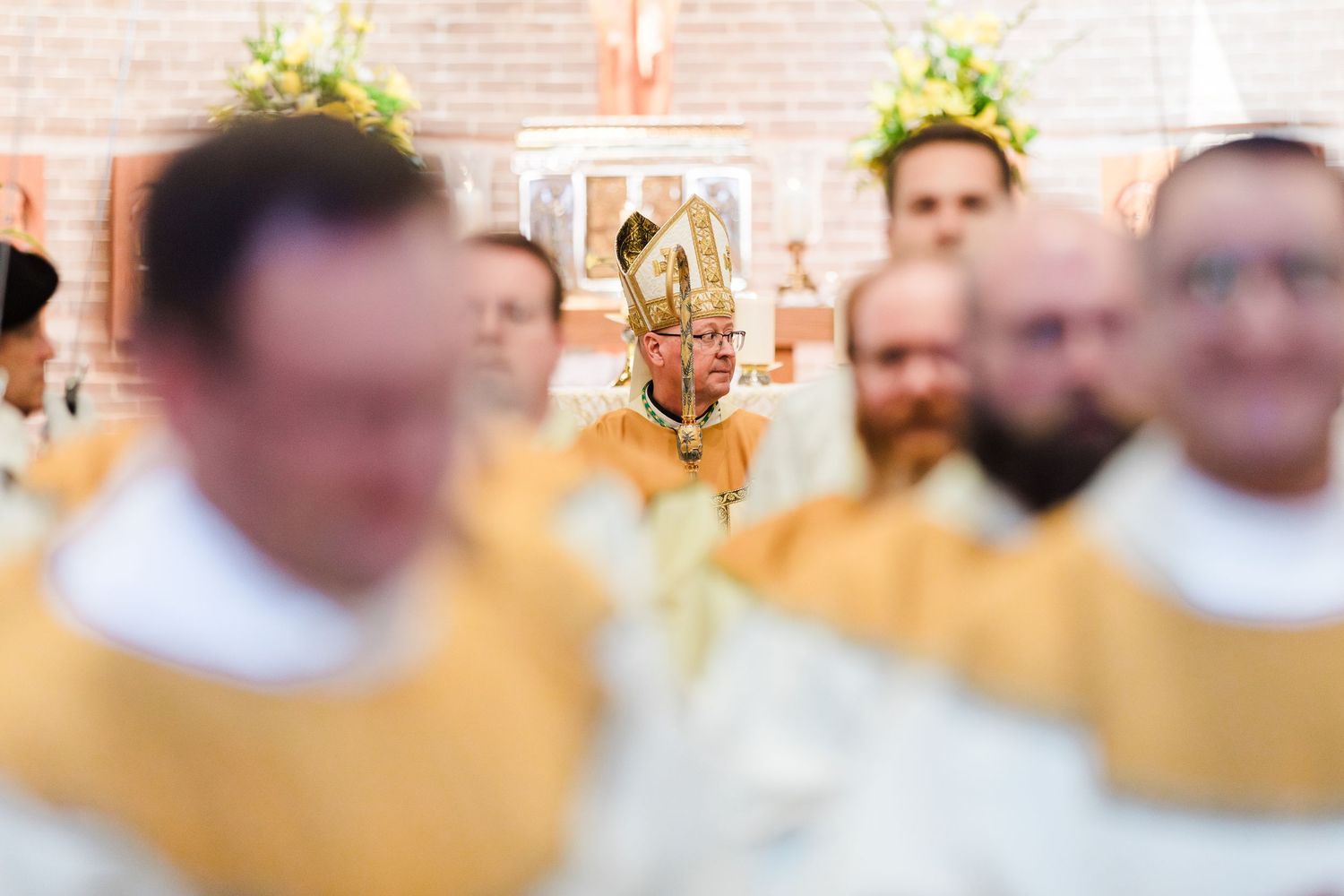 Catholic bishop in golden vestments presides over an ordination ceremony with priests in yellow robes in a brick church.